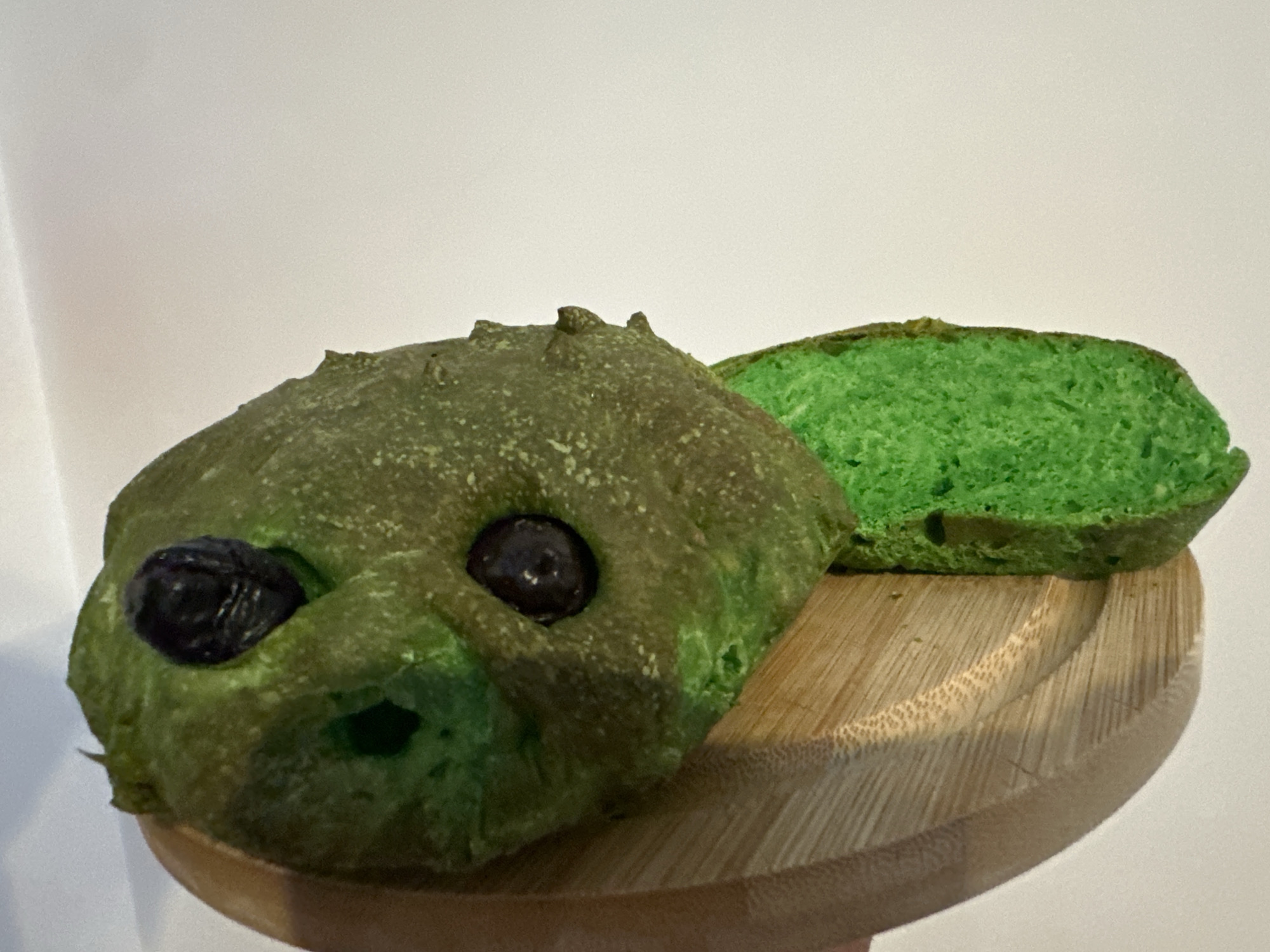 A green, alien-shaped bread bun with two dark olive eyes sits on a wooden circular cutting board. A slice of the same green bread, showing its airy interior, is placed behind the bun. The background is a plain, light-colored surface. The image does not provide any information about a location in Moortown, Ireland.
