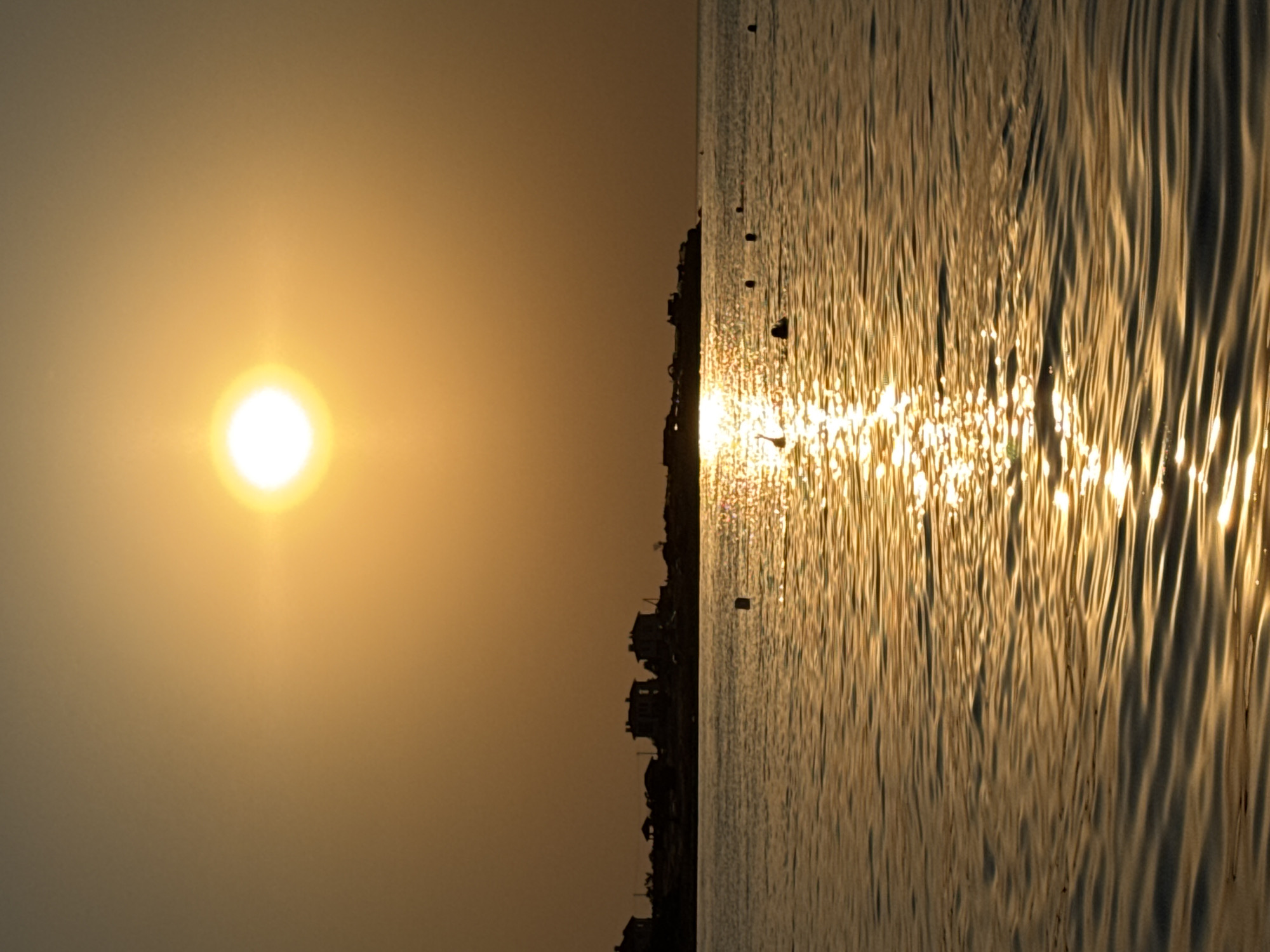 This is a photograph of a sunset over the sea, possibly in Sevastopol, Ukraine. The sun is a bright, golden orb near the top center of the image, casting a warm glow across the sky and water. In the distance, silhouetted against the sunset, is a coastline with several buildings. The sea is calm, with gentle waves reflecting the sunlight. There are several buoys visible in the water, as well as a few figures swimming. The overall impression is one of tranquility and natural beauty.