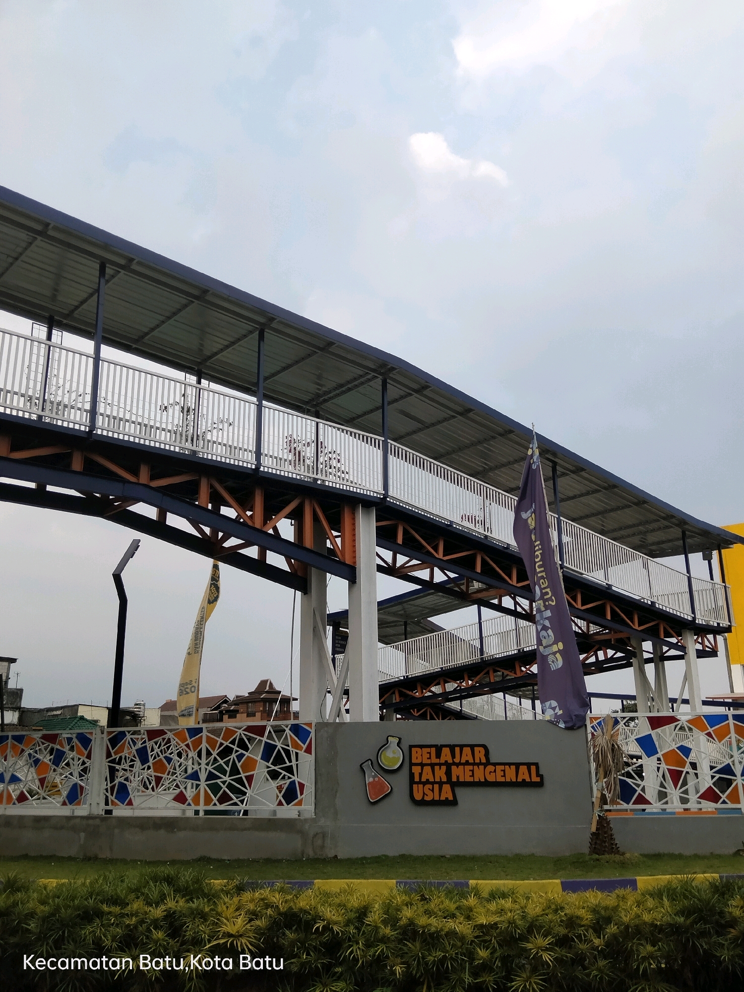 The image shows a pedestrian bridge with a metal roof and white railings, supported by blue and orange steel beams. In front of the bridge is a gray wall with a colorful geometric pattern. To the left of the wall, there is a sign with two stylized flasks and text that reads "BELAJAR TAK MENGENAL USIA". A purple flag with white text is planted near the wall. In the background, several buildings are visible. The bottom of the image has text indicating the location: "Kecamatan Batu, Kota Batu", which translates to Batu District, Batu City in Indonesia. The sky is overcast.