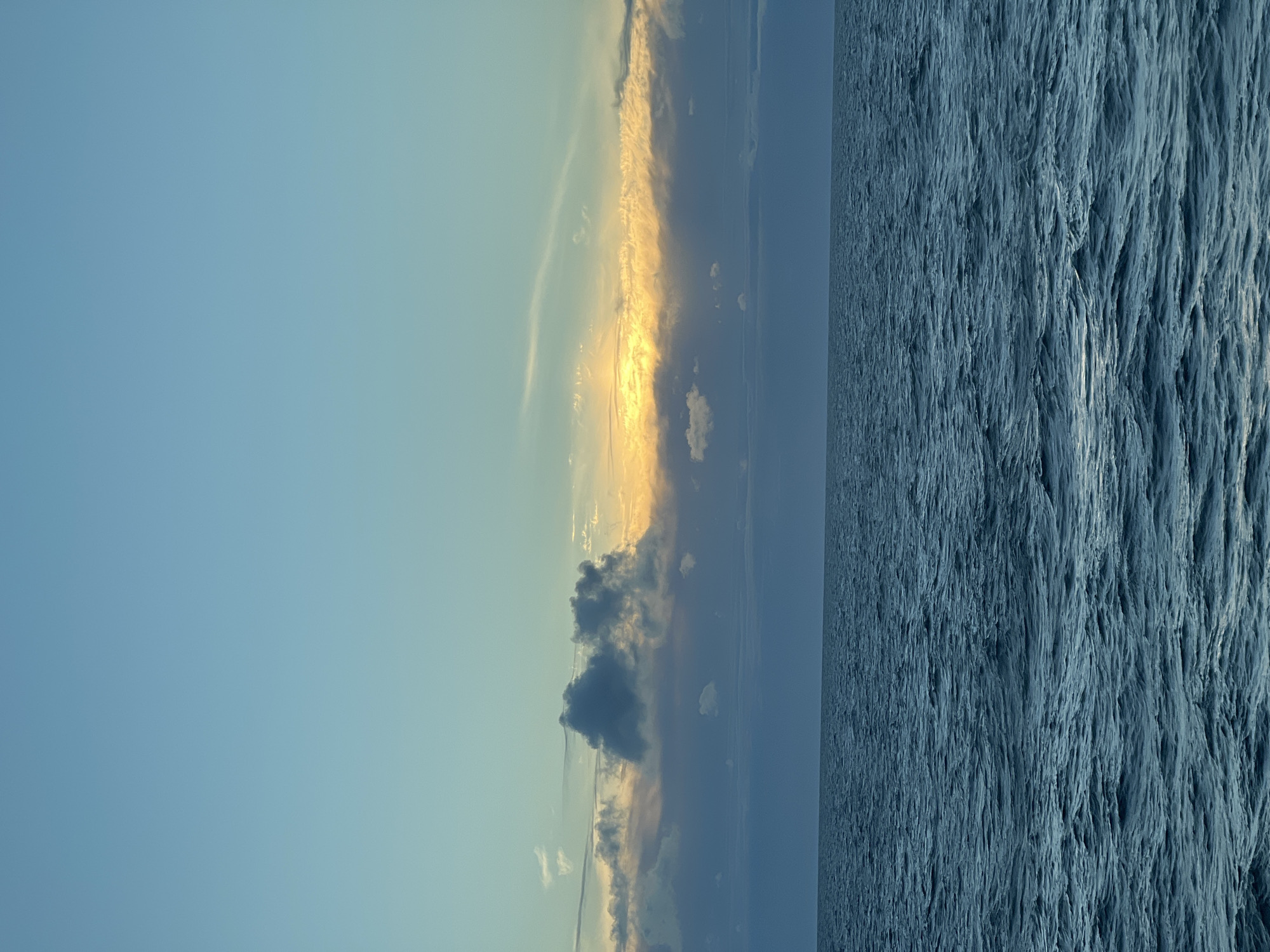 An eye-level, medium shot captures a tranquil expanse of the Baltic Sea under a serene, partially clouded sky at dusk. The sky transitions from a pale blue at the top to warmer hues of yellow and orange near the horizon, where sunlight breaks through a band of clouds. Darker, billowy clouds are scattered across the sky, with some casting shadows on the water below. The sea is a deep, rich blue, with gentle, undulating waves creating a textured surface. The horizon line is clearly defined, separating the vastness of the water from the ethereal sky. The scene evokes a sense of peace and natural beauty, with no people or man-made structures visible. The location is identified as the Baltic Sea near Poland.