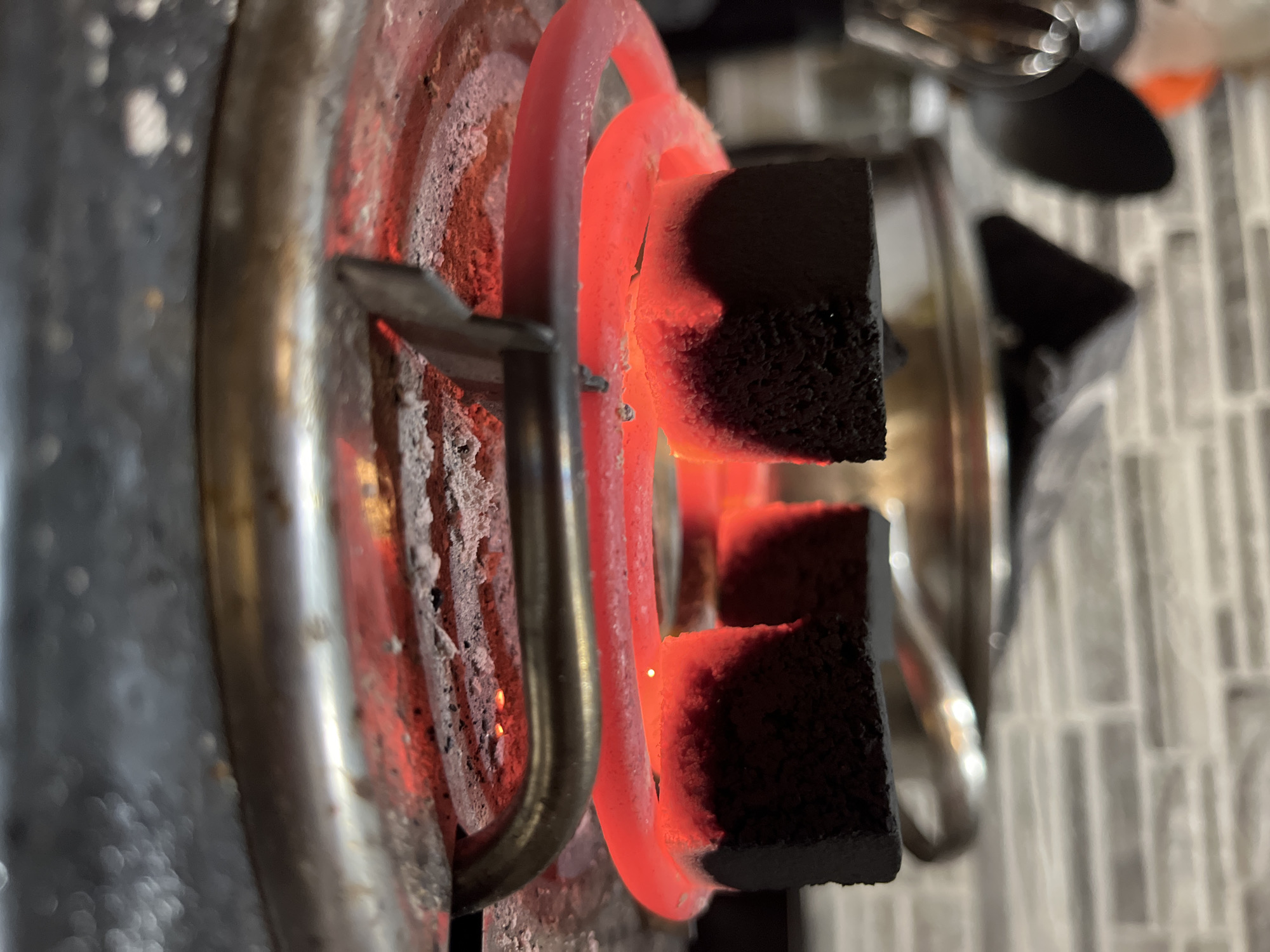 The media file captures a close-up, high-angle shot of two dark, square-shaped charcoal briquettes being heated on a glowing red electric stove coil. The coil itself is incandescent, emitting a bright red and orange light that illuminates the surrounding area and the charcoal. White, dusty residue, possibly ash or burnt food, is visible on the coil and the metallic support frame beneath it.
The charcoal briquettes, with their porous and rough texture, are placed directly on the hot coil, appearing to be in the process of ignition or already glowing with heat themselves. A tiny, bright white spark or hot spot is discernible where the coil touches one of the charcoal pieces.
In the blurred background, parts of a shiny, metallic object, likely a pot or kettle, are visible. Behind this, a light-colored, possibly white or grey, tiled surface with a grid pattern can be seen, suggesting an indoor kitchen environment. The primary light source is the intense glow from the stove coil, casting strong contrasts and shadows. No people or text are visible in the image. The scene conveys an activity of preparing coals, possibly for shisha or a similar purpose, within a kitchen setting in what is described as Chaikovsky, Russia.