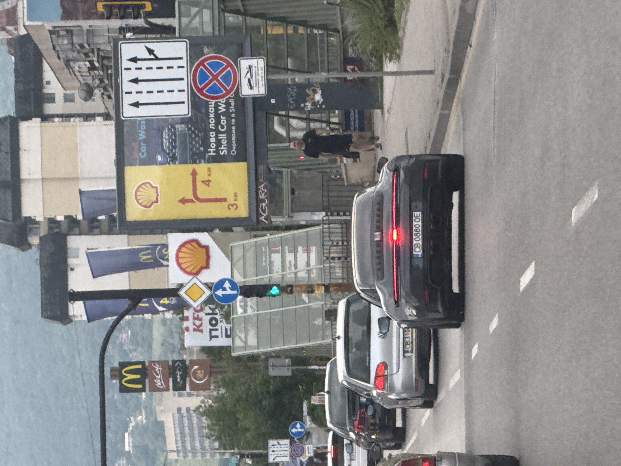 The image captures a street scene in Sofia, Bulgaria, specifically near the Pliska Hotel intersection on Tsarigradsko Shose boulevard, evidenced by the combination of prominent commercial signs, the architecture of the surrounding buildings, and the "CB" prefix on vehicle license plates.
The scene depicts multiple vehicles halted in traffic on an asphalt road with white dashed lane markings. In the immediate foreground, the rear of a dark grey Porsche (license plate "CB 0880 OE") is visible, with its red brake lights illuminated. Behind it, a silver Volkswagen Golf (license plate "CB 310") is stopped. Further back, a dark sedan is also stationary. To the far left, a portion of another vehicle is present.
On the right side of the frame, a man in a black t-shirt and black shorts walks on the sidewalk, carrying a black bag in his left hand. The sidewalk is bordered by a metal railing and sparse green vegetation. A utility pole stands on the sidewalk, featuring a "no stopping/parking" sign (red cross on a blue circle) and a supplementary sign indicating vehicle towing for violations.
Dominating the mid-ground and background are numerous signs and urban structures. Commercial signs include a prominent McDonald's "M" logo and "McCafé" sign, a KFC logo with Cyrillic text (partially obscured), multiple Shell logos (red and yellow scallop shells), and a brown round sign displaying "24". A large billboard advertises a "Nova lokacia Shell Car Wash" (New location Shell Car Wash) and provides directional information with distances: 3 km to the left and 4 km to the right. A blue vertical banner with an "M" (McDonald's) is also visible.
Traffic signs include a yellow diamond (priority road ahead), a blue circular sign with a white arrow pointing right (mandatory right turn), and a white rectangular sign depicting three straight-through lanes and one right-turn only lane merging from the right. A green traffic light is visible above the street.
Behind the array of signs, a modern pedestrian underpass entrance/exit structure made of glass and metal is visible. In the background, multi-story buildings of varying architectural styles are present, with a large, light-colored building featuring a dark roof particularly noticeable. Distant hills or mountains, covered in vegetation, are visible under an overcast sky.