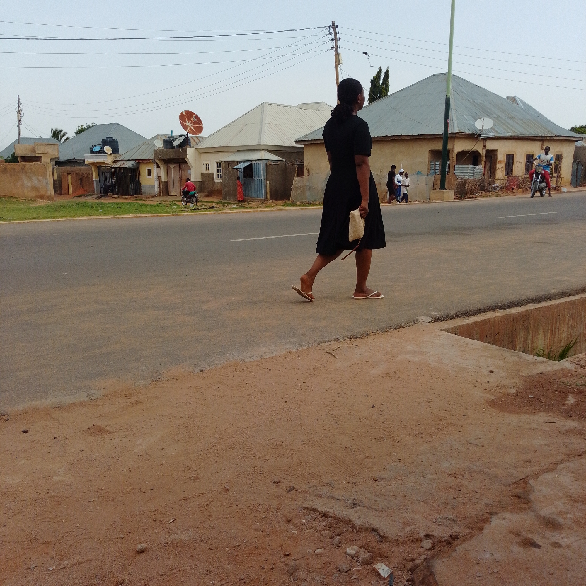 The image captures a street scene in Gombe, Nigeria, with a focus on a woman walking on the right side of the frame. She is wearing a black dress and carrying a light-colored bag in her right hand. The woman is walking away from the camera, and her left foot is slightly raised, indicating movement. She is wearing flip-flops on both feet.
In the background, several buildings with corrugated metal roofs are visible, characteristic of architecture in the region. A prominent satellite dish is mounted on one of the buildings. A paved road runs horizontally across the image, with a rough, unpaved area in the foreground.
Further down the road, to the left, a man on a red motorcycle is visible. To the right, a man on a blue and white motorcycle is also present. Several other individuals are walking in the background near the buildings. The sky is overcast, suggesting either early morning or late afternoon lighting. The environment appears to be an urban or semi-urban area, likely a residential or commercial street within Gombe.