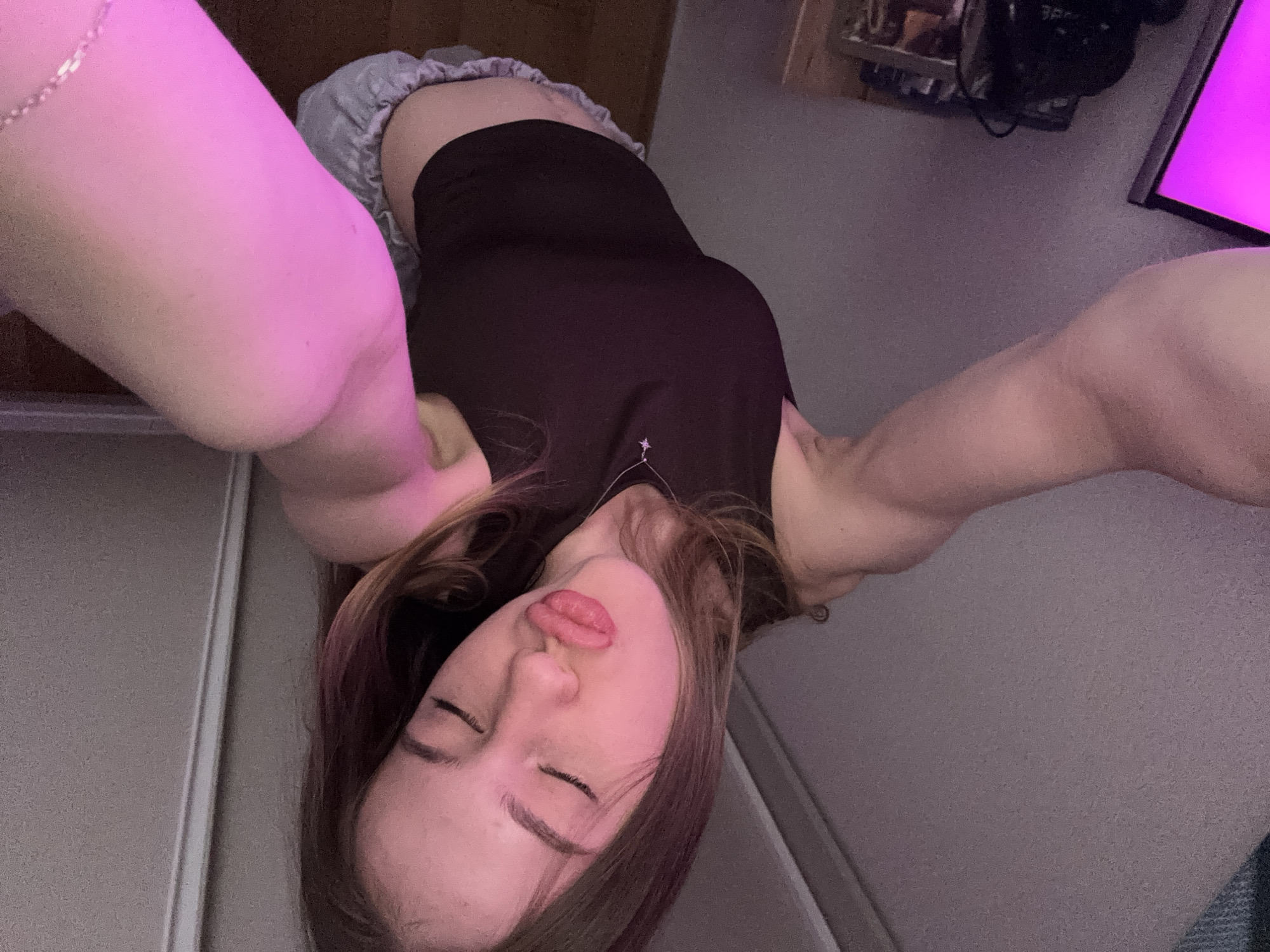 A young woman is captured in a close-up, high-angle selfie, appearing to be lying down or leaning back with her arms extended upwards, holding the camera. The image is oriented with her head at the bottom and her body extending upwards. She has long brown hair, with some strands falling around her face. Her eyes are closed, and her lips are pursed in a playful or kiss-like expression, highlighted with a reddish-pink color. She wears a dark-colored tank top or bodysuit, and light-colored shorts or underwear with a ruffled waistband are visible. A delicate necklace with a small pendant rests on her chest.
The scene is an indoor setting, likely a bedroom or personal space, characterized by dim lighting. A vibrant pink or magenta light illuminates her left arm and leg, casting a soft glow on her skin and the surrounding area. In the background, to the left, a light-colored wall or door frame is visible. To the right, various objects are partially obscured by shadow, including what appears to be a shelf with items and a screen displaying a bright magenta light. The overall atmosphere is intimate and relaxed, suggesting it's evening or nighttime. There is no visible text in the image. The location context provided is Novoe Devyatkino, Russia.