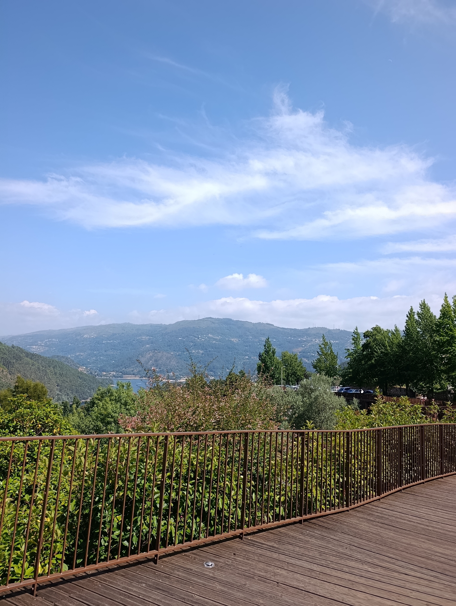 The image depicts a scenic viewpoint overlooking a valley with a body of water and rolling hills in the distance. In the foreground, there is a wooden walkway with a weathered metal railing. Lush green trees and bushes line the edge of the walkway and extend into the valley. The sky is a clear blue with scattered white clouds. The overall impression is one of a peaceful, natural landscape, possibly a park or a viewpoint in a mountainous region. The specific location in Terras de Bouro, Portugal, is suggested by the architecture and vegetation, characteristic of the region.