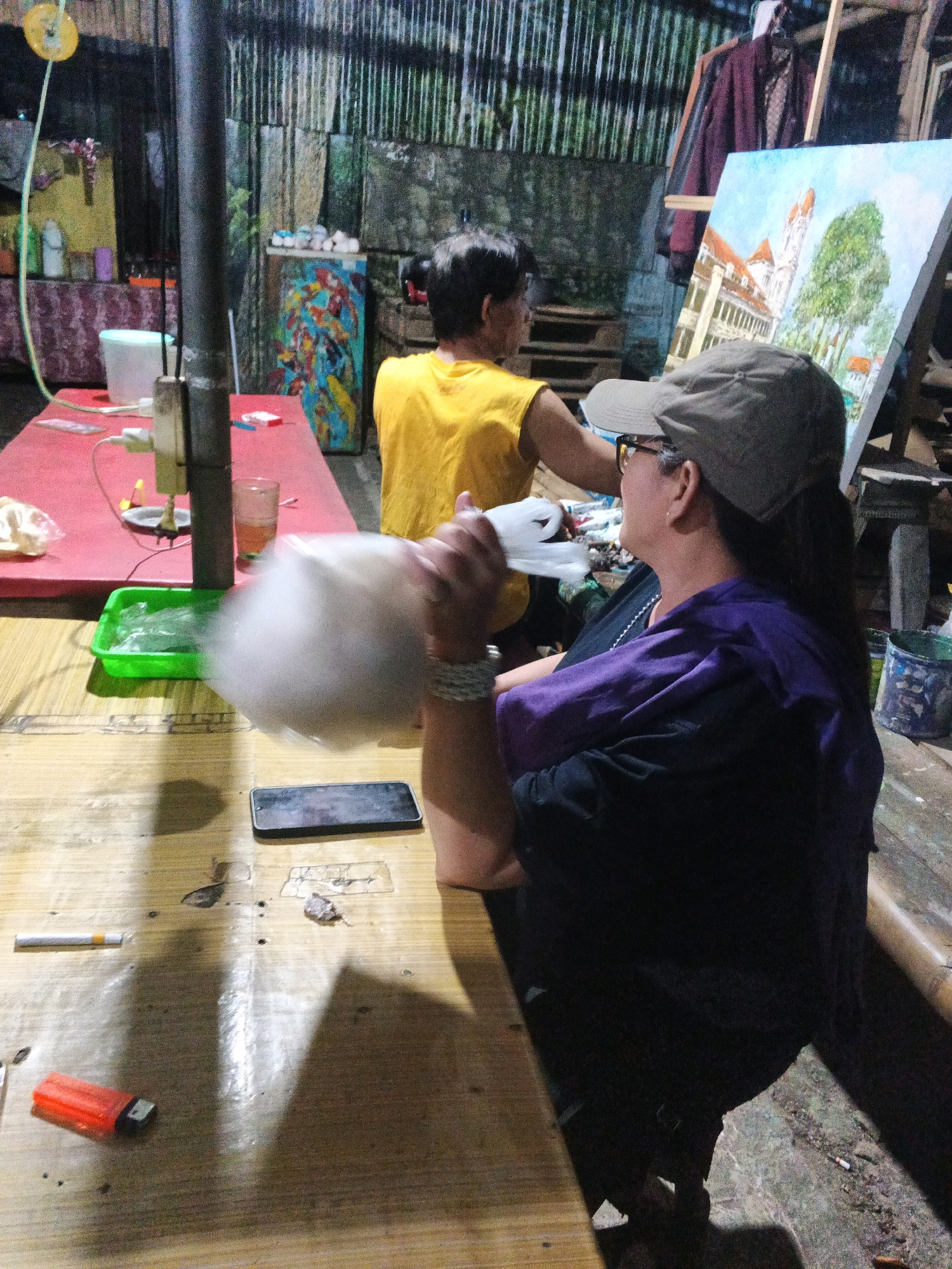 The image depicts an outdoor scene in what appears to be a workshop or informal art studio in Ungaran, Indonesia. In the foreground, a person wearing a purple top and a grey cap is seated at a wooden table, holding a white object that is blurred due to motion. On the table are a smartphone, a cigarette, and an orange lighter. To the left, a red table holds a plastic container with ice and a glass of amber liquid. In the background, another person in a yellow shirt is visible. An easel with a painting of a building and trees is set up, and various art supplies and materials are scattered around. The environment suggests a casual, working atmosphere with a backdrop of textured walls and hanging garments.
