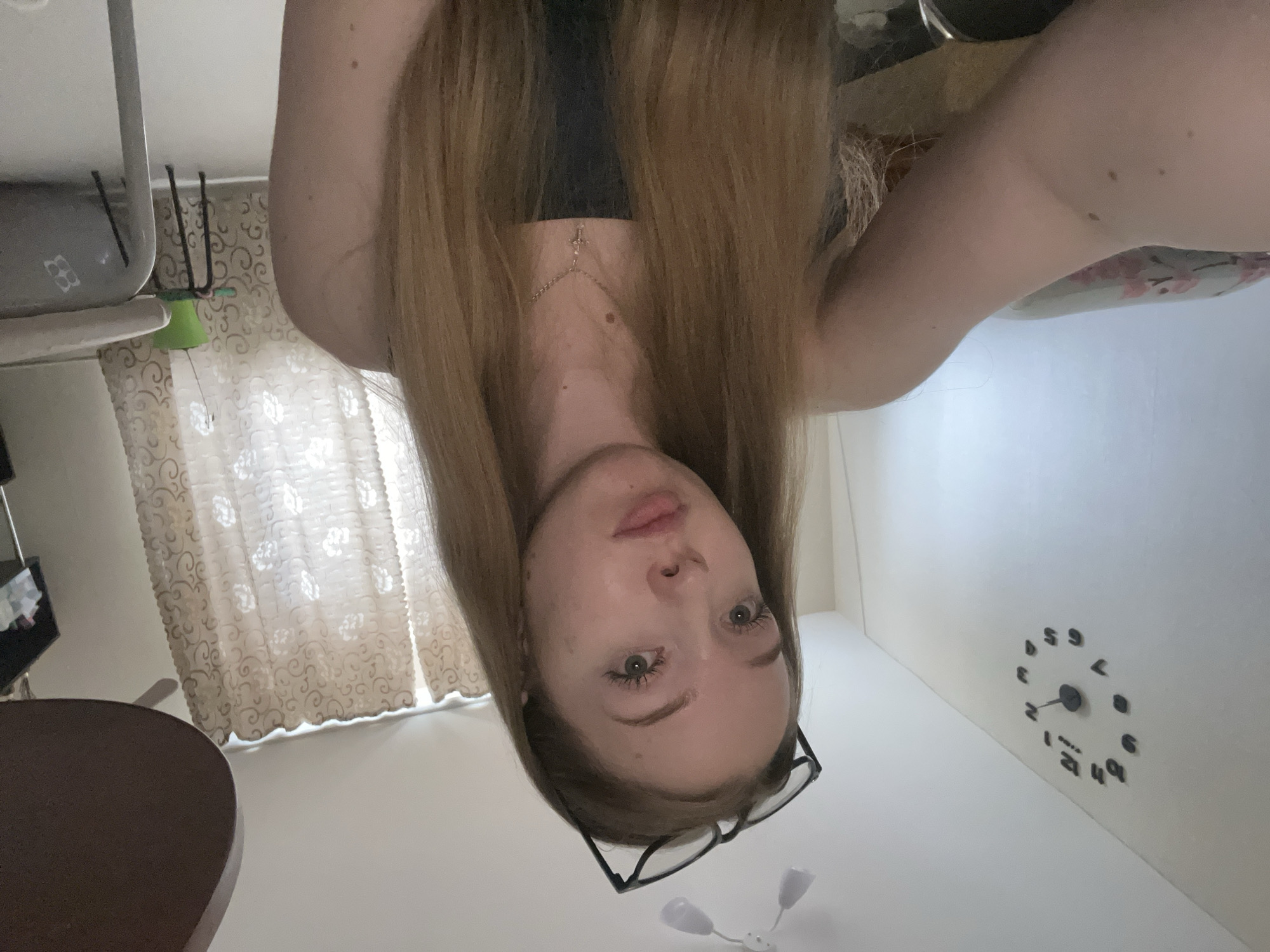 An indoor selfie captures a young woman looking directly up at the camera from an overhead perspective. She has long, straight, light brown hair that frames her face, falling around her shoulders. Her complexion is fair, and she has light-colored eyes. She wears a simple black top and a delicate chain necklace with a small cross pendant. A pair of black-framed glasses rests on her forehead or pushed back into her hair. Her expression is neutral and direct.
In the background, to the left, sheer, light-colored curtains with a subtle patterned design are visible, indicating a window. Below the window, a dark brown, possibly wooden, round table is partially seen. Further left and slightly above the table, a light-colored piece of furniture, possibly a shelf or part of a chair, is present, with dark, slender objects (perhaps chair legs or a stand) behind it.
To the right of the woman, a plain white wall extends. A minimalist wall clock with individual black numbers (1 through 12) placed around a central mechanism is affixed to this wall. Below her head, against the white wall, there's a small white object with two small, light-colored protrusions, possibly a small light fixture or decoration. The lighting suggests daytime, likely from the window.