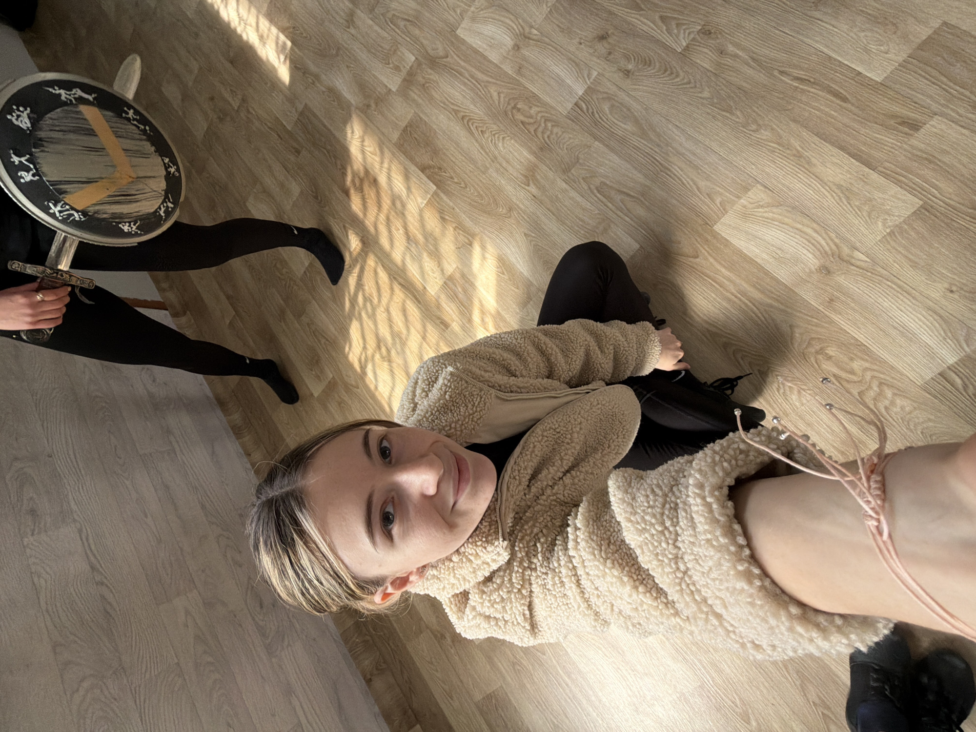A medium-shot photograph depicts a woman taking a selfie from a top-down perspective, situated in a room with light-colored wood laminate flooring. The woman is wearing a beige sherpa jacket over a black top and black leggings. She is smiling at the camera.
To the left of the woman, a person is holding a sword and a round shield with symbols and numbers on it. The shield appears to be leaning against a white chair. This person is wearing black tights and shoes.
The location is indoors, with sunlight streaming in at an angle. There is no evidence to suggest this photograph was taken in Plovdiv, Bulgaria.
