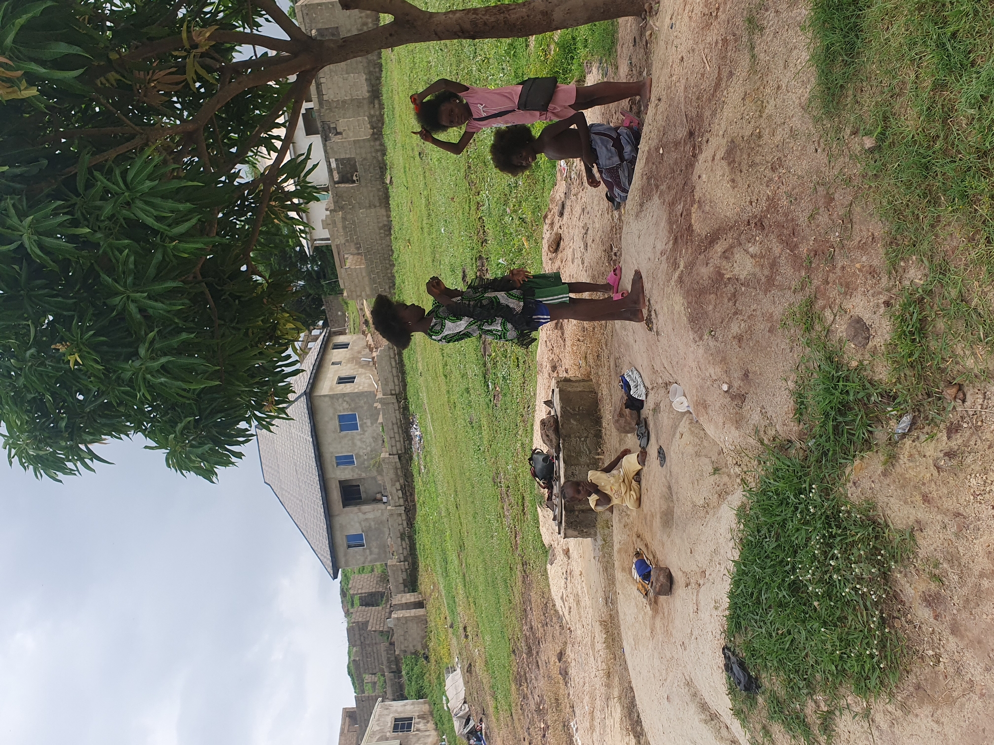 This is a medium shot photograph taken outdoors, likely in Lokoja, Nigeria, under an overcast sky. The scene depicts several children playing near a tree. In the foreground, a child in a yellow outfit is sitting on a sandy mound. To the right, three other children are standing. One child, wearing a pink shirt and shorts, is holding an object in their hair. Another child in a patterned dress and green shorts stands nearby, appearing to be looking at something in their hand. A third child, shirtless and wearing patterned shorts, is kneeling.
Behind the children, the environment consists of a grassy slope dotted with partially constructed buildings made of concrete blocks. A finished house with a gray roof and blue-rimmed windows is visible on the upper portion of the slope. The ground in the immediate vicinity of the children is sandy and uneven, with some rocks and scattered objects like shoes and bags. A large tree with lush green leaves is prominent in the right side of the frame, providing shade. The overall impression is one of a rural or developing urban setting.