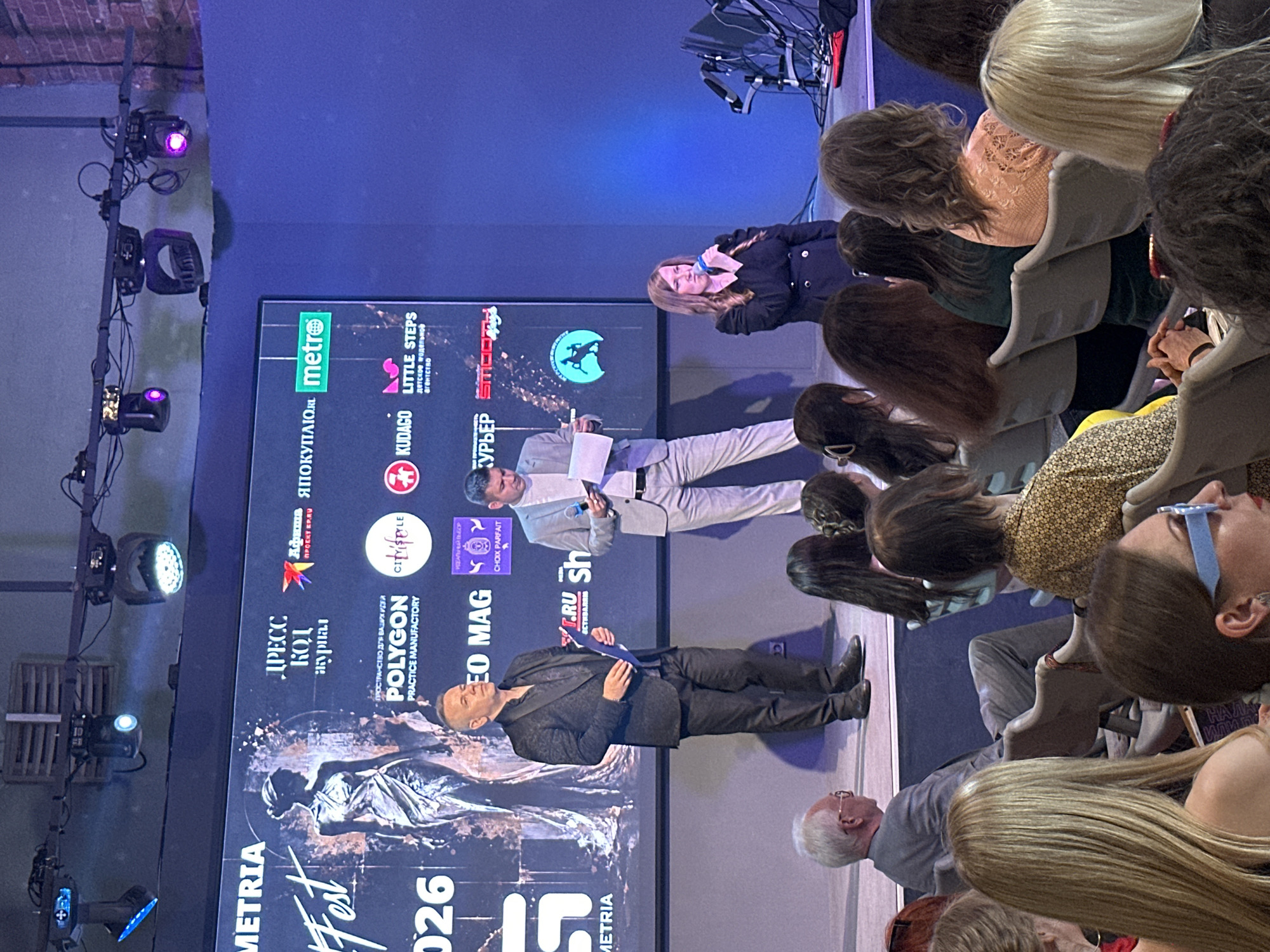 The image depicts an indoor event with a stage and an audience. On the stage, three individuals are visible. The leftmost person is a man in a dark, patterned suit, holding a few sheets of paper. To his right, another man in a light grey suit is holding a microphone and looking down at papers. The rightmost person on stage is a woman with blonde hair, wearing a black jacket, speaking into a microphone. Behind them is a large screen displaying logos and text. In the foreground, an audience is seated, facing the stage, with their backs primarily to the camera. The environment appears to be a conference hall or event space, with stage lighting visible overhead.