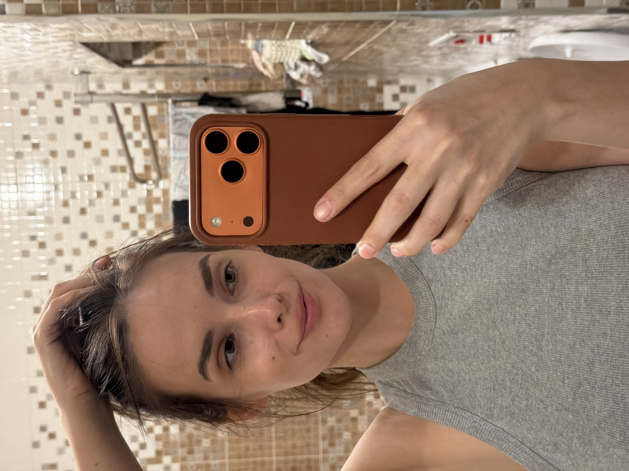 A young woman with brown hair and brown eyes is taking a selfie in a bathroom. She is wearing a grey tank top and is holding an orange phone in her right hand. Her left hand is in her hair. The bathroom has tiled walls with a checkered pattern of brown, beige, and white squares. A towel rack is visible in the background, with towels hanging on it. The overall lighting is dim, typical of an indoor environment.
