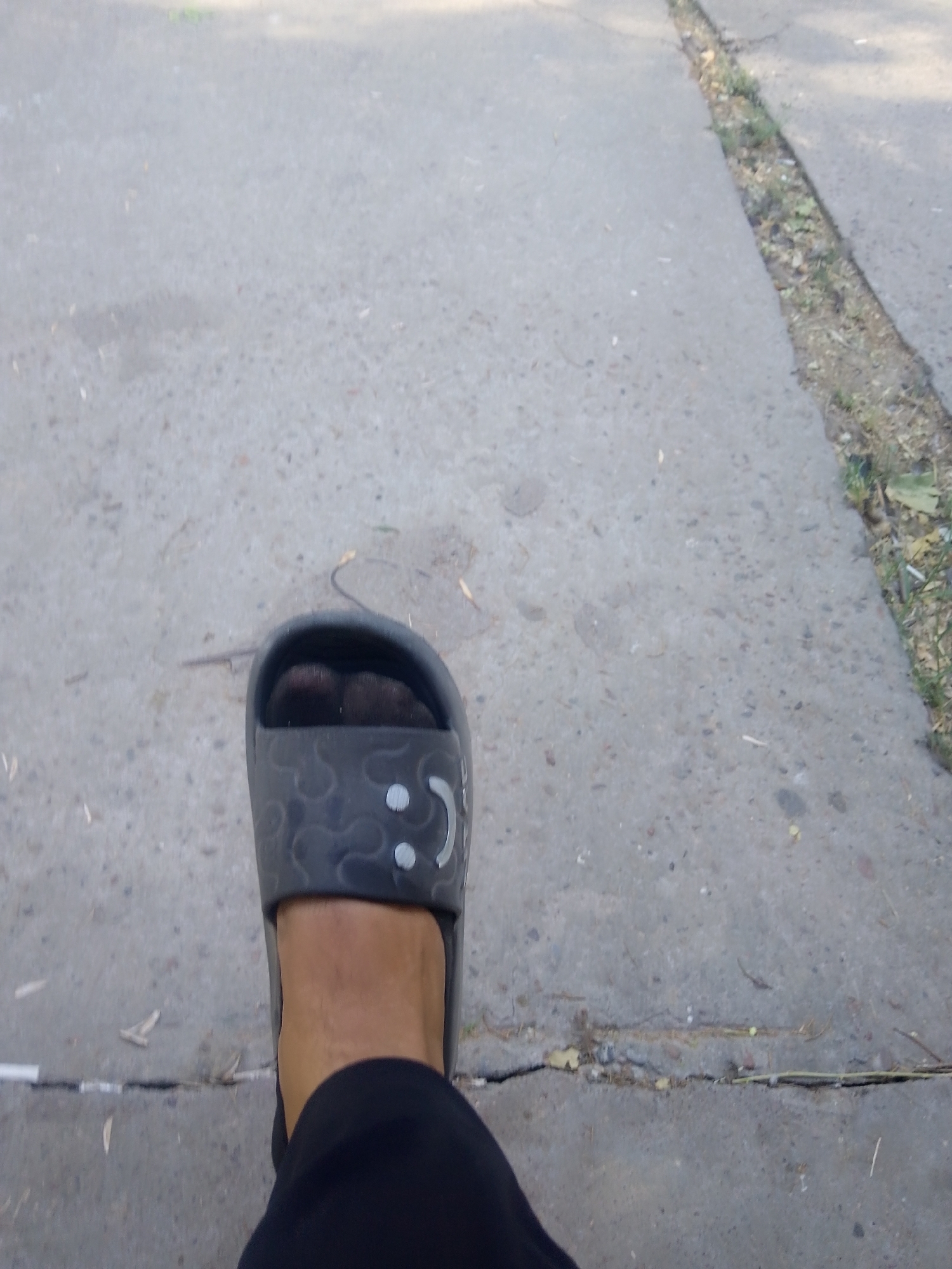 The image shows a close-up, point-of-view shot of a person's left foot wearing a black slide sandal. The sandal has a pattern of swirling lines and small circular indentations, with two slightly raised circles on the strap resembling eyes. The foot is clad in dark pants that obscure the ankle. The sandal is positioned on a cracked concrete surface, possibly a sidewalk or path. To the right of the concrete, there is a narrow strip of dirt and sparse vegetation. The lighting suggests an outdoor, daytime setting. The location is identified as Абай ауылдық округі, Kazakhstan.