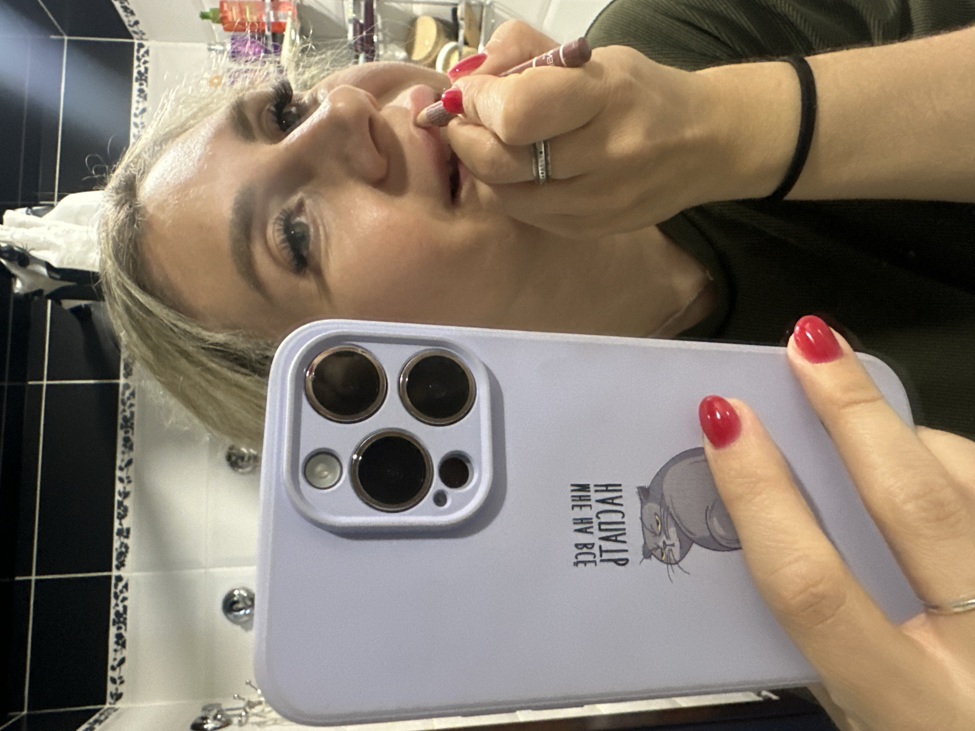 A blonde woman is applying makeup to her lips in front of a mirror. She is holding a light purple smartphone in her left hand, which is reflecting her face. She is wearing a dark green top and has bright red fingernail polish. The background appears to be a bathroom with black and white tiled walls. There are some toiletries visible on a shelf in the background. The image does not provide any information that would indicate the location in the city of Novoaltaysk, Russia.