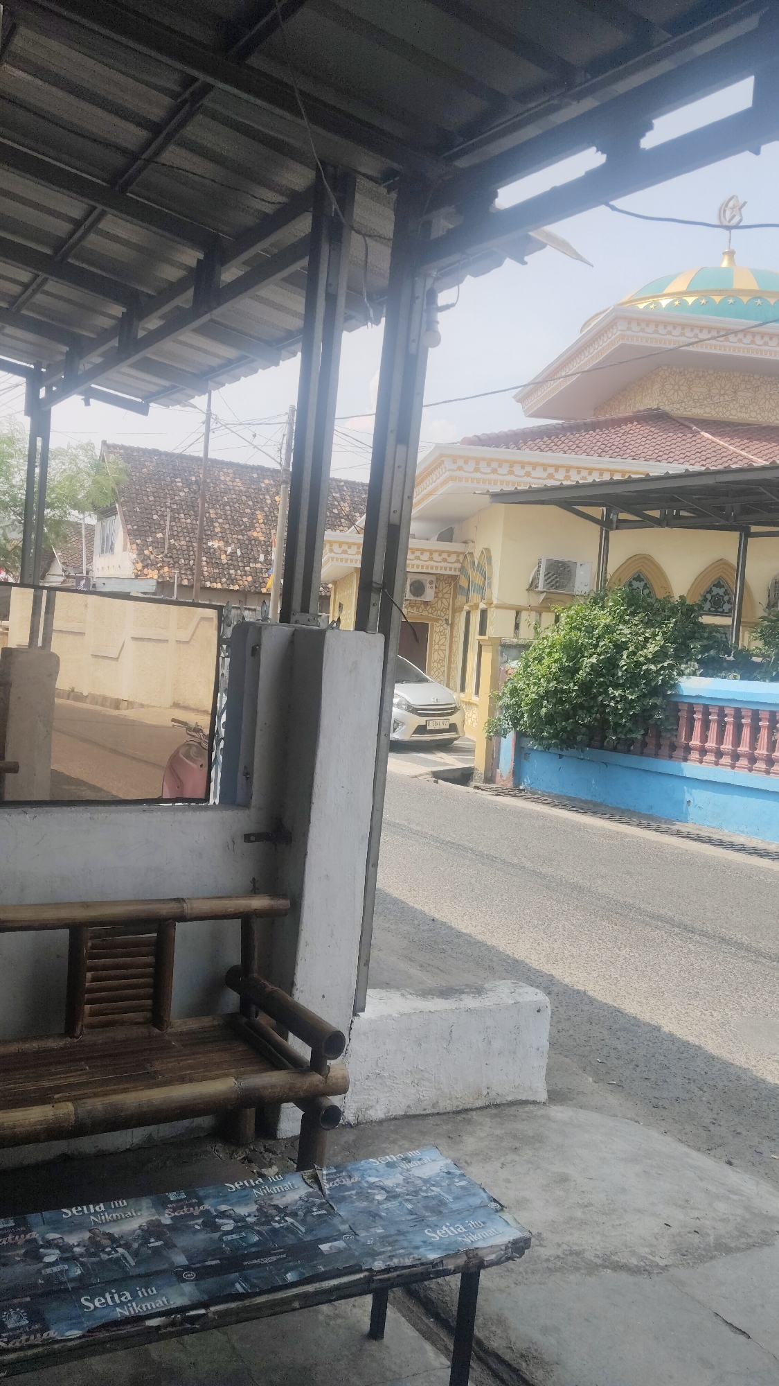 This is a photograph taken from a covered outdoor area looking towards a street in Bandar Lampung, Indonesia. In the foreground, a bamboo bench sits next to a table covered with a poster featuring motorcycles and text in Indonesian. To the right of the bench, a concrete curb separates the seating area from the street. The street itself is paved and appears to be a typical urban road.
Further down the street, a light-colored car is parked. On the right side of the street, there is a mosque with a prominent green and yellow dome topped with a crescent moon. The mosque has yellow walls with decorative arched windows. Adjacent to the mosque, a dense green bush grows. A pink and blue fence runs along the edge of the property.
In the background, traditional Indonesian houses with tiled roofs are visible, suggesting a residential neighborhood. The sky is bright and clear, indicating a sunny day. The overall scene depicts a common urban landscape in Indonesia, with a focus on the street view and the presence of a mosque.