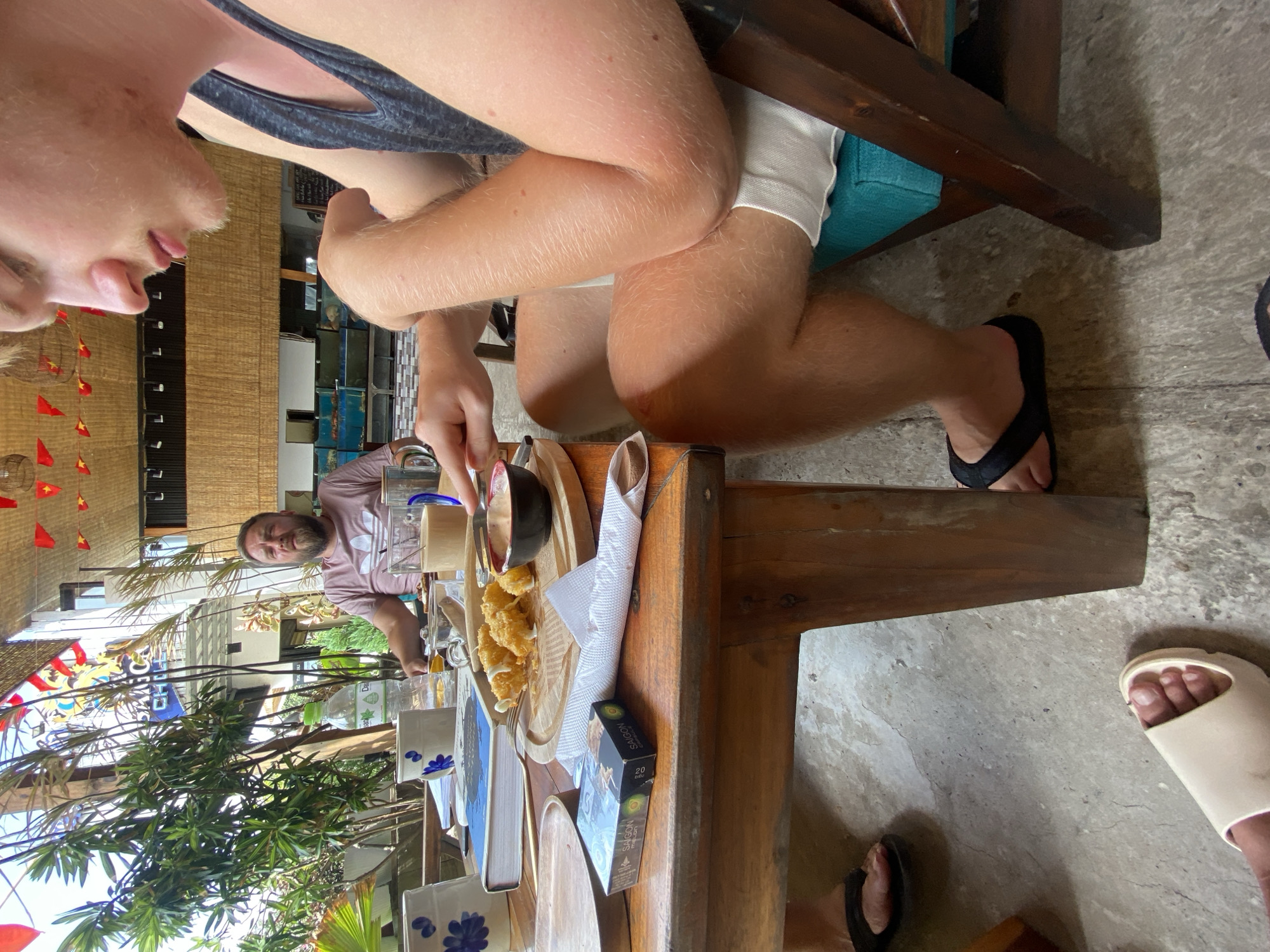 A daytime image depicts a semi-outdoor restaurant or cafe scene in Phu Thuy Ward, Vietnam. Three individuals are seated at a wooden table on a cracked concrete floor.
In the foreground, a young adult male with fair skin and light stubble wears a dark grey tank top and white shorts. He sits on a wooden chair with a turquoise cushion. His right hand holds a fork, poised above a small black bowl containing a reddish-pink dipping sauce. The bowl is on a round wooden board next to several fried, possibly cheese-coated, food items. His feet, clad in black flip-flops, are visible on the concrete.
To the mid-left, an adult male with a beard and fair skin wears a light pink t-shirt with an Adidas logo. He is seated at the table, looking directly forward. A clear glass bottle of "Dalat" water and a clear glass mug containing a light brown beverage are near him.
Additional items on the wooden table include two white mugs with blue floral patterns, a blue-covered book, and a green pack of "SAIGON MELON" cigarettes. On the far-left foreground, a foot wearing a light beige slide sandal is partially visible.
The background features lush green foliage, buildings, and a partially visible "SAIGON CAFE" sign. Red and yellow triangular flags (likely Vietnamese national flags) are strung overhead, and woven bamboo panels are visible as part of the decor.