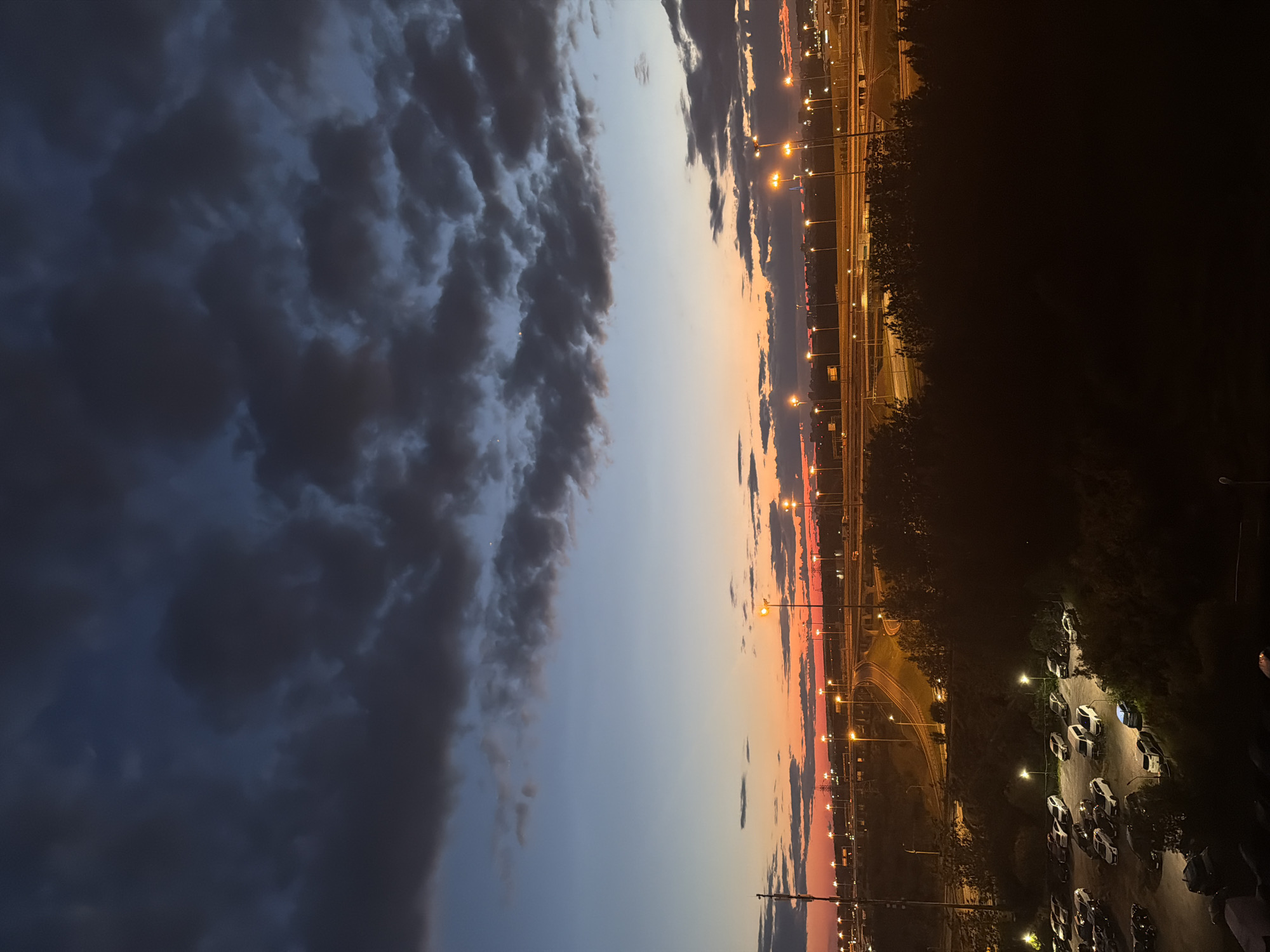 This is a photograph of a city skyline at dusk. The sky is filled with dark, dramatic clouds, with bands of orange and pink light near the horizon. Below the sky, a highway interchange is visible, with numerous streetlights illuminating the road and surrounding areas. In the foreground, a parking lot with several cars is visible, as well as a dense line of trees. The overall impression is one of an urban landscape at the end of the day, with the artificial lights of the city beginning to dominate the natural twilight.