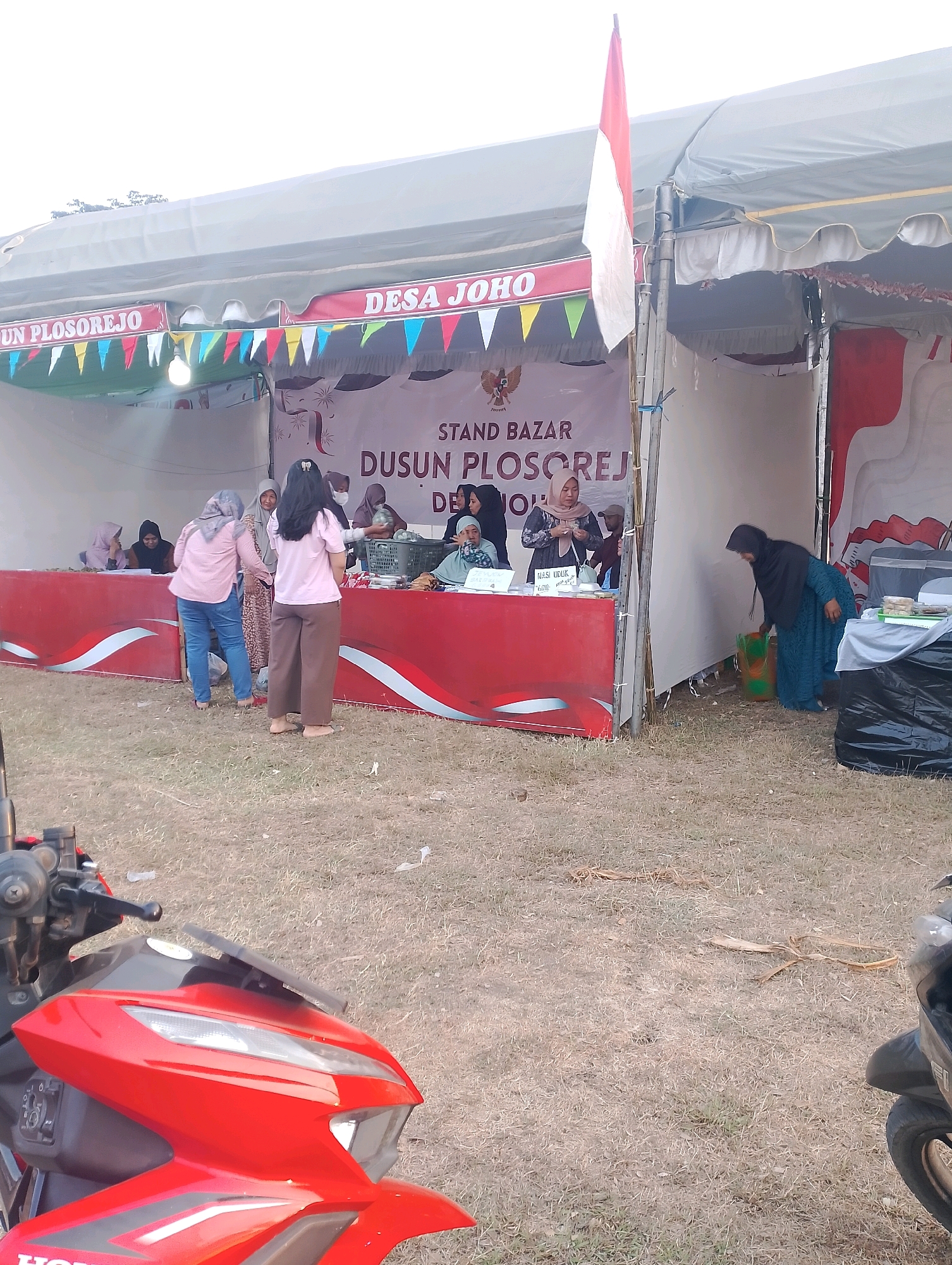 A street market stall in Joho, Indonesia, is set up under a large white tent. The stall is decorated with red and white trim and colorful triangular flags. A large banner reads "DUSUN PLOSOREJO" and "DESA JOHO." Several people are gathered at the stall, some interacting with each other and others looking at merchandise. One woman in a blue dress is bending down to pick up a green bag. In the foreground, the front of a red motorcycle is visible. The ground is covered with dry grass and a few scattered leaves.
