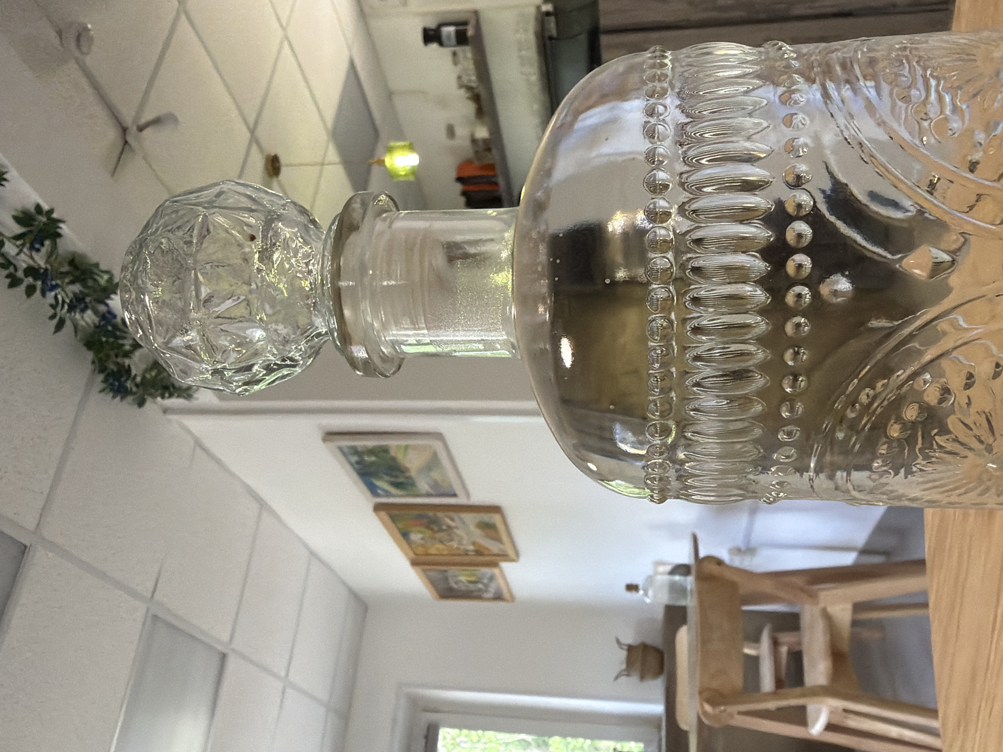 The image shows a clear glass decanter with an ornate stopper and beaded detailing. It is resting on a light-colored wooden surface. In the background, there's a white ceiling with recessed lighting and hanging framed artwork. A window provides a view to the outside, showing greenery. The scene depicts an interior, likely a residential kitchen or dining area. No specific location details in Chaikovsky, Russia, can be determined from the image.