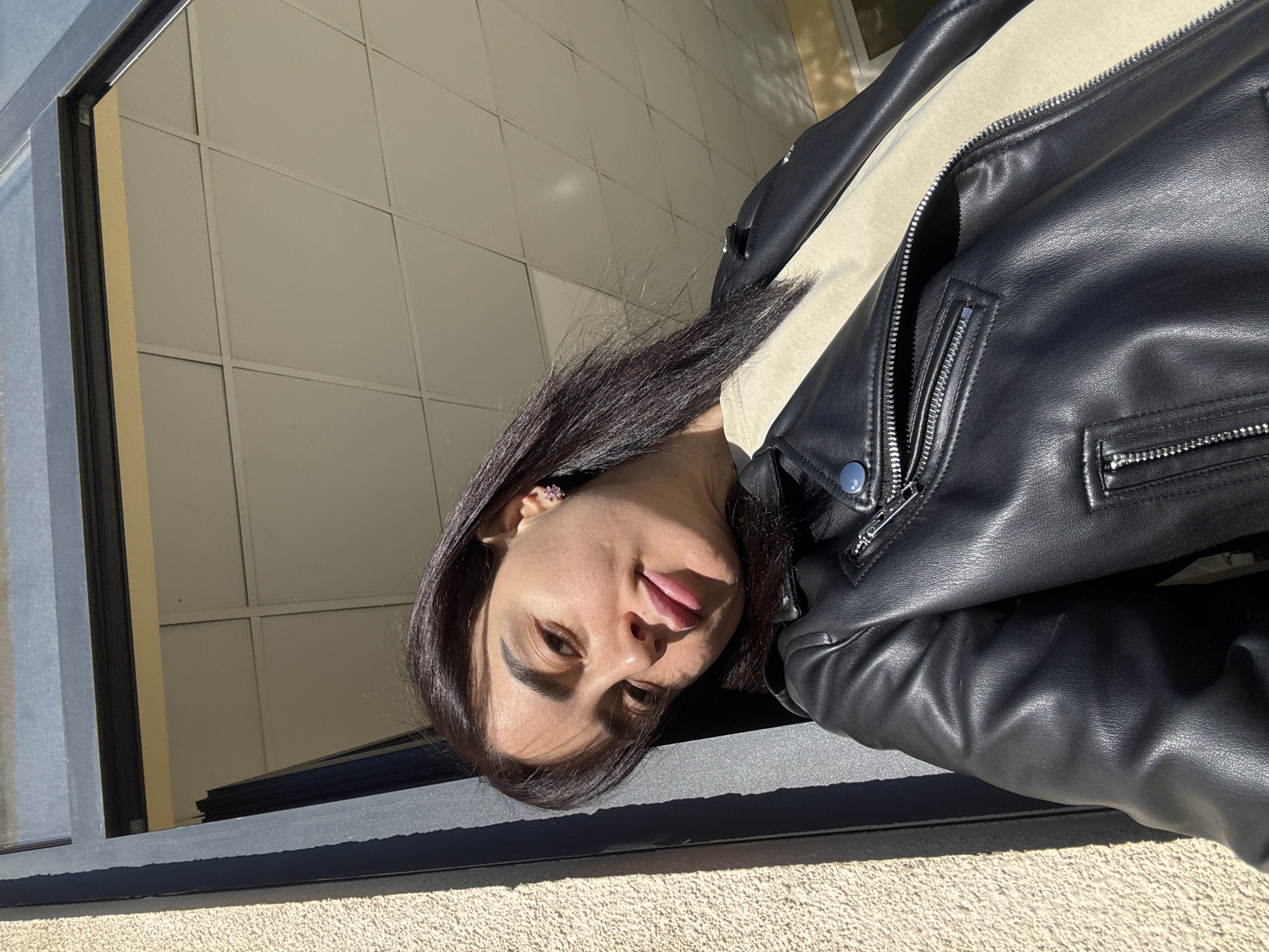 A woman wearing a black leather jacket is posing for a selfie in front of a building. The building appears to have a textured exterior and metal-framed windows. The location is Tbilisi, Georgia.