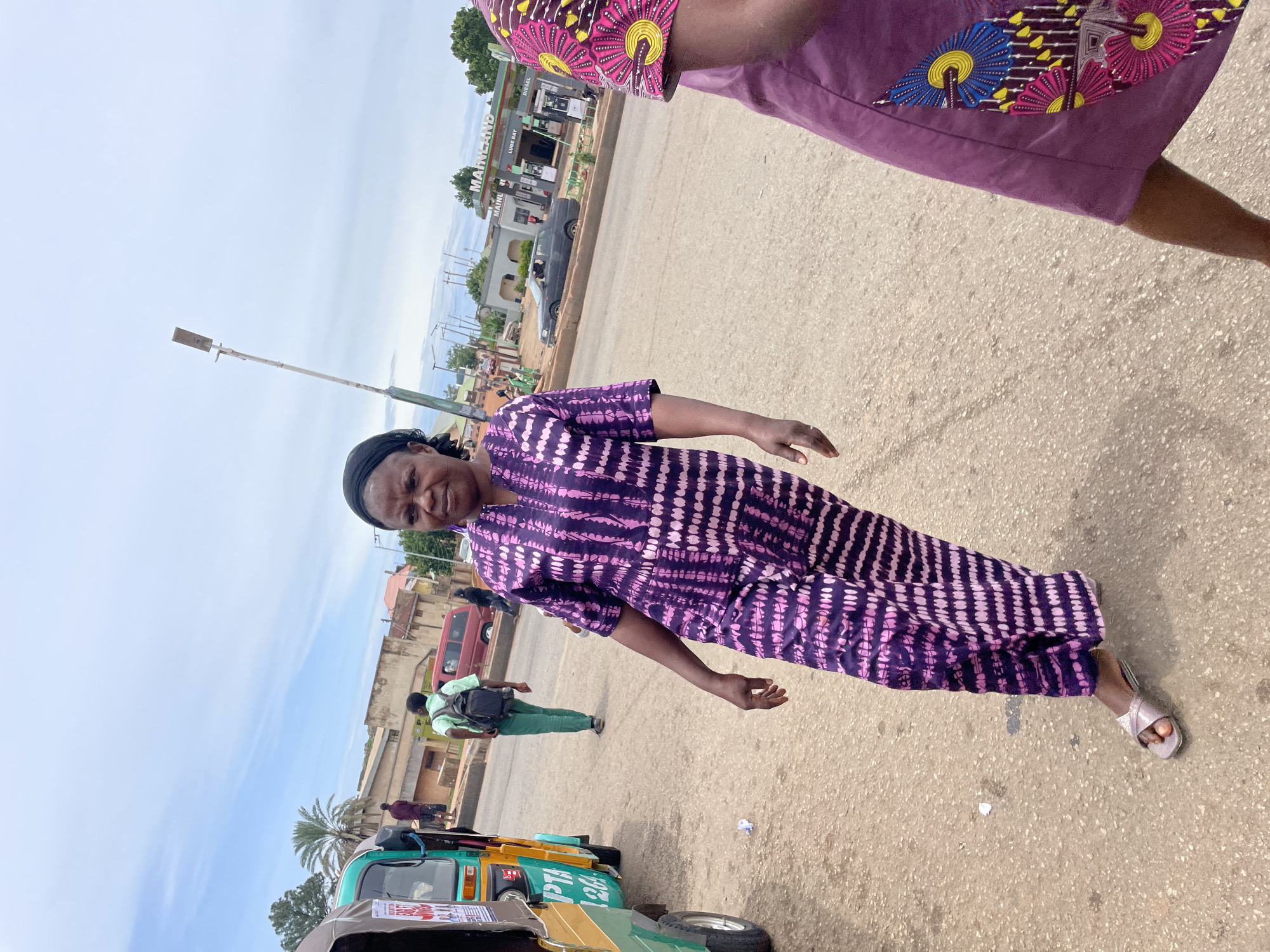 An outdoor medium shot captures a woman walking on a paved road in Gura Top, Nigeria. She is wearing a purple and pink tie-dye patterned outfit and a black headscarf. Her right arm is bent at the elbow, and her left arm is partially visible. To her left, a green and yellow auto-rickshaw is parked on the side of the road. In the background, a red van is visible, as well as a gas station with a sign that reads "MAINLAND LUBE BAY DIESEL". The sky is partly cloudy.