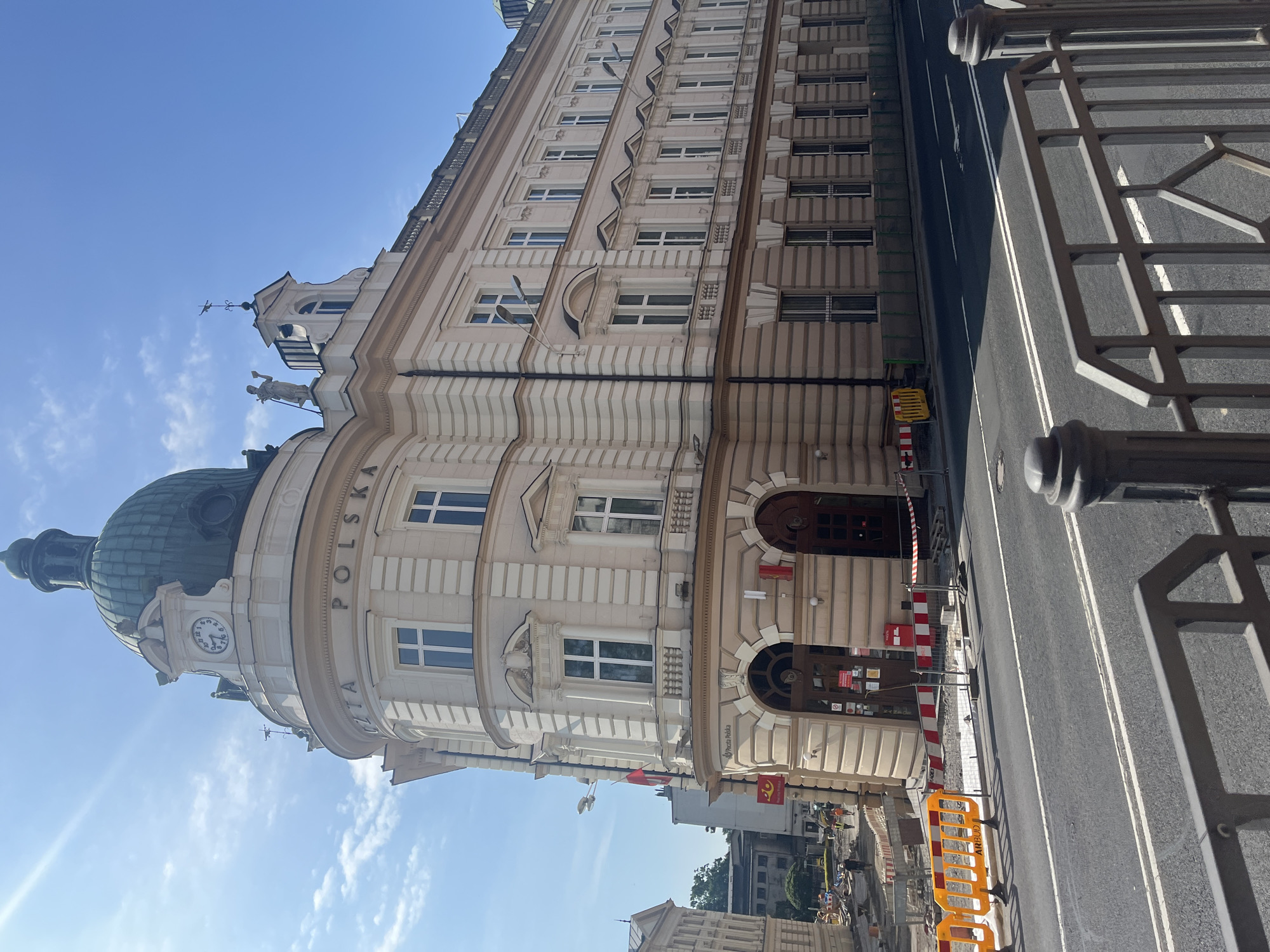 A three-story, light-beige building with a green dome is partially visible on a sunny day. The building features numerous windows, decorative molding, and a clock on its facade. A sign above the entrance reads "Poczta Polska". In front of the building, there is a street with a black asphalt surface and white markings. Construction barriers and orange fences are present along the sidewalk, indicating ongoing work. A few people are visible in the distance, near the construction area. The sky is blue with scattered clouds.