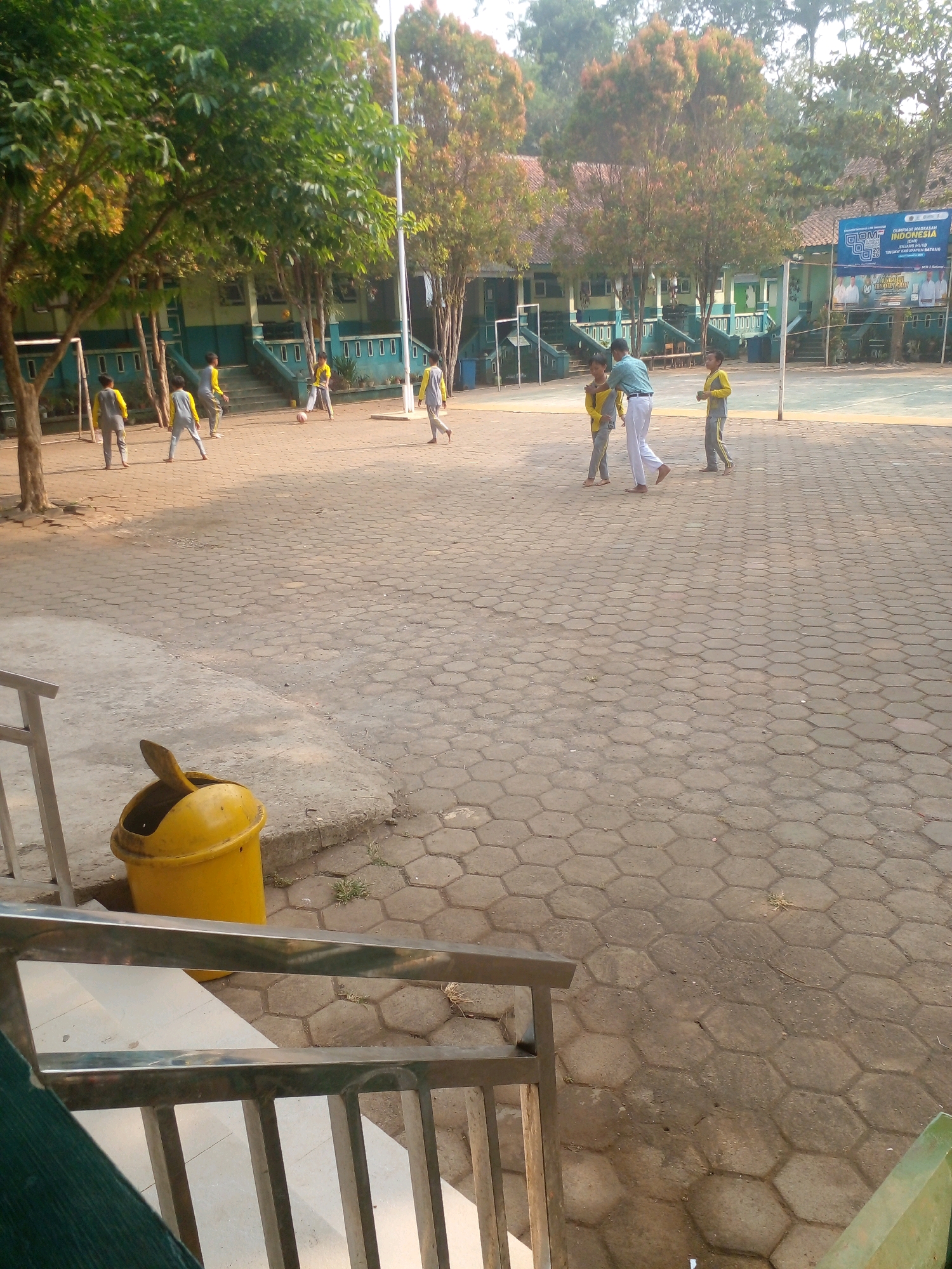 A group of young boys, wearing yellow and gray athletic wear, are playing soccer in the courtyard of a school in Banyuputih, Indonesia. One boy is actively kicking the ball, while others are running and positioned on the field. A teacher, dressed in a light blue shirt and white pants, appears to be observing or interacting with the students. The school building, with its distinctive green and white facade, serves as a backdrop. The courtyard is paved with hexagonal tiles, and a yellow trash can is visible in the foreground. Trees with lush green foliage surround the school grounds. The scene is bathed in the warm light of what appears to be late afternoon sun.