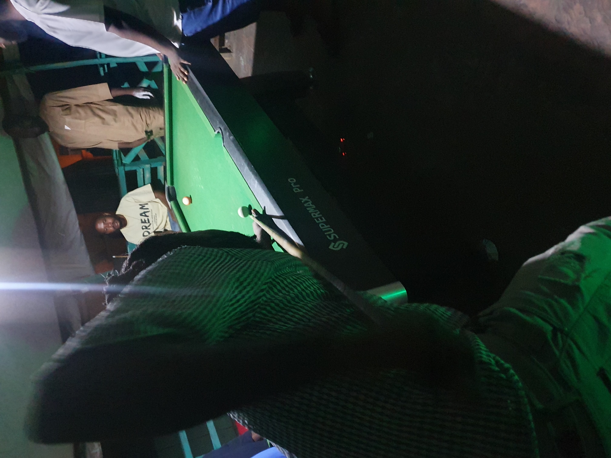 The image depicts a group of men playing billiards in what appears to be an outdoor, dimly lit setting, possibly a bar or a recreational area. The scene is in Lokoja, Nigeria. A pool table with green felt and black edging is central to the image, with several billiard balls visible. One person, wearing a light-colored shirt with "DREAM" printed on it, is seated near the table, seemingly observing. Another man, dressed in a brown kaftan, stands behind the table, his hands clasped. To the right, another man, wearing a white t-shirt and blue jeans, is partially visible, with his hands resting on the table. A fourth individual, whose face is not clearly visible due to the angle and lighting, is actively playing, holding a cue stick. The overall atmosphere is casual and social, with a spotlight or bright light source casting strong highlights and shadows. The background suggests an open-air structure with supporting beams and a draped canopy.