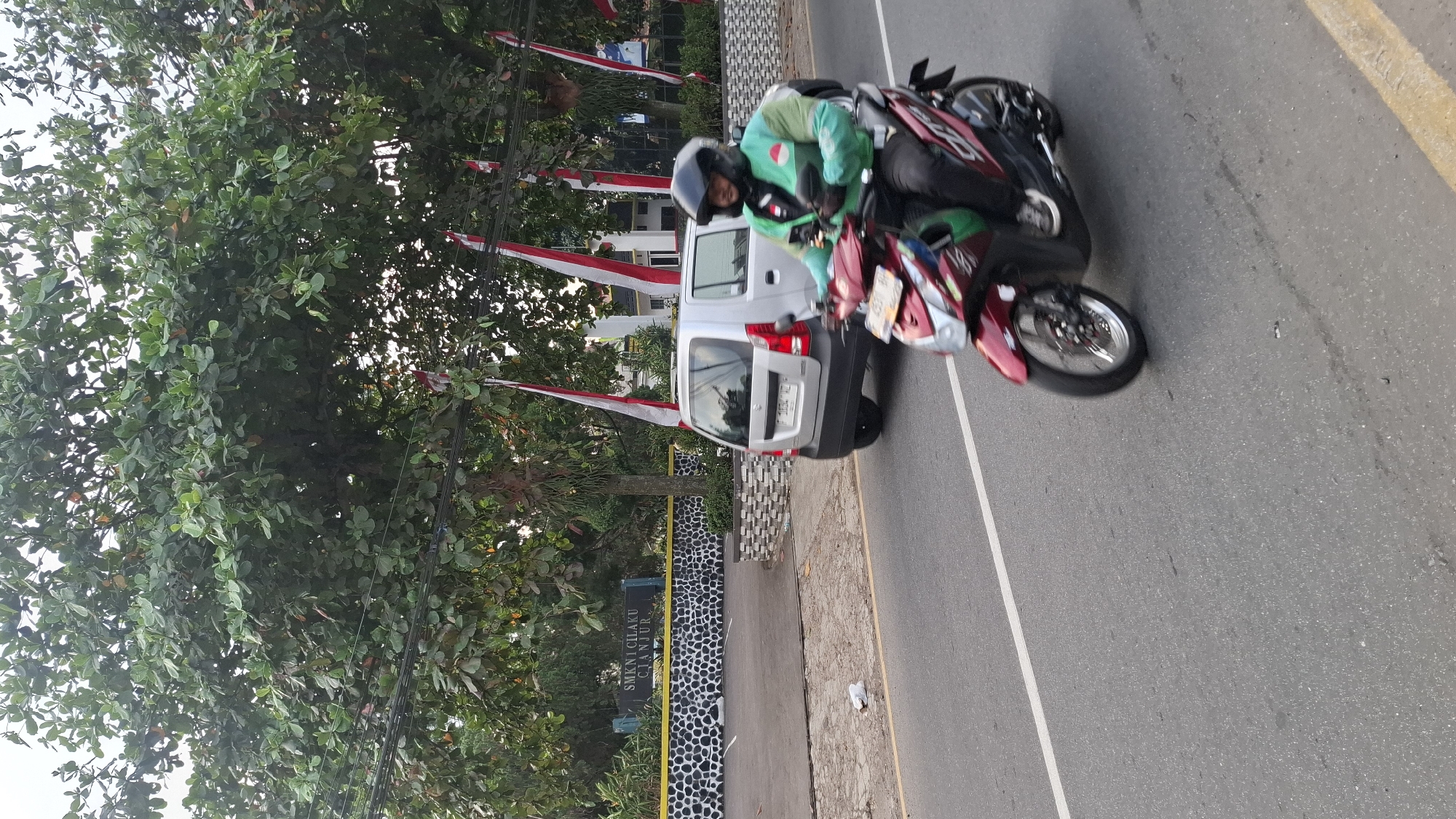 A man wearing a green jacket and a black helmet rides a red scooter on a paved road in Cilaku, Indonesia. He is seen from the side, moving forward. Behind him, a silver car is parked on the side of the road, near a building with a black and white patterned wall. The building has a sign that reads "SMK N 1 CILAKU CIANJUR". Large trees with green leaves are present on the left side of the image, and red and white flags are displayed on poles. The road has a white line marking the lane. The sky is overcast.