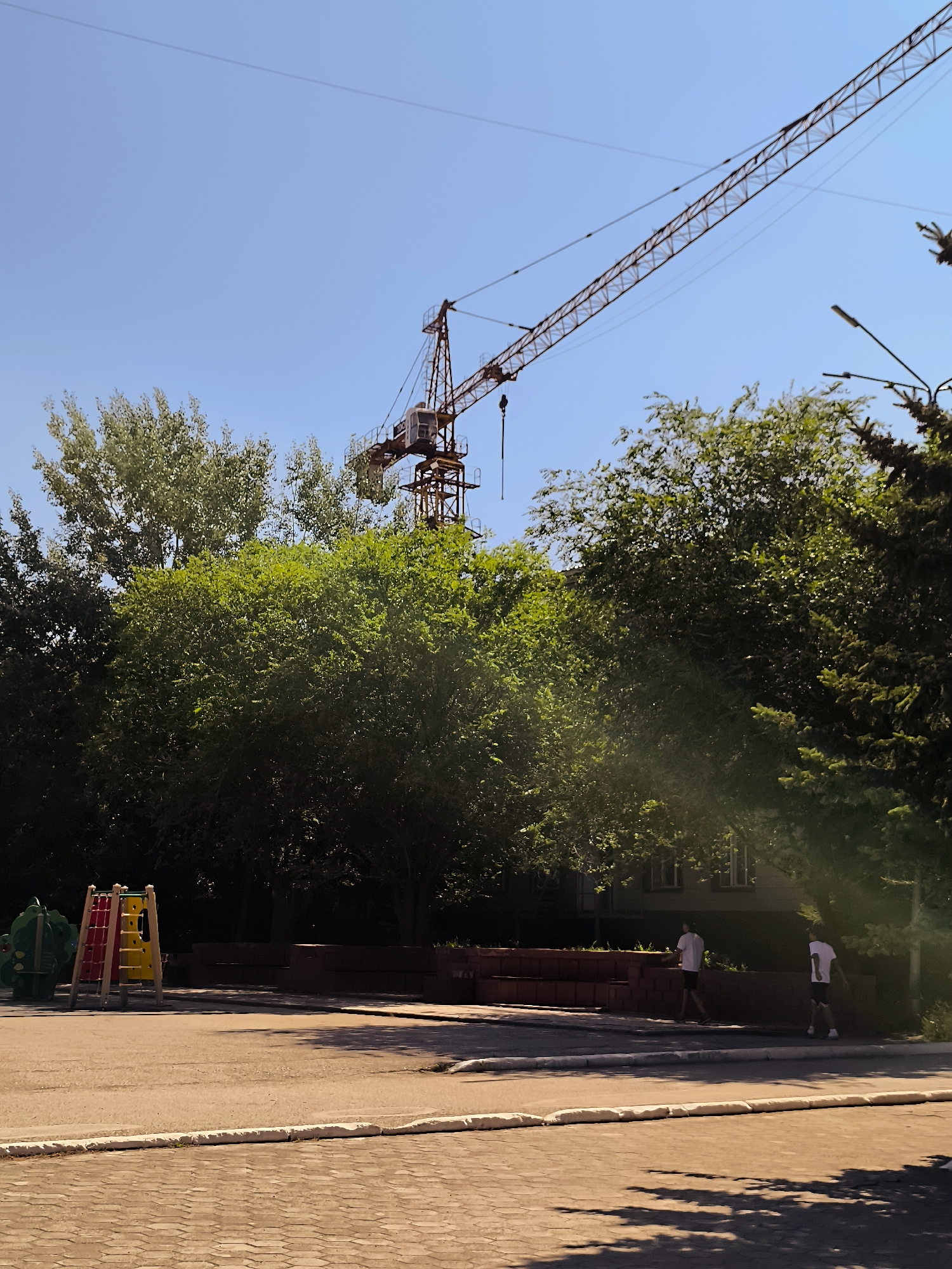 The image depicts an outdoor scene on a clear day, likely in Qarağandı city administration, Kazakhstan. In the background, a large construction crane with a red and yellow boom is prominent against a bright blue sky, extending diagonally across the upper portion of the frame. Below the crane, a dense canopy of green trees partially obscures a building. In the foreground, a playground area is visible on the left with brightly colored play structures, including a slide and climbing wall. To the right, two individuals wearing white t-shirts and dark shorts are walking on a paved pathway. The ground in the foreground is covered with patterned paving stones.