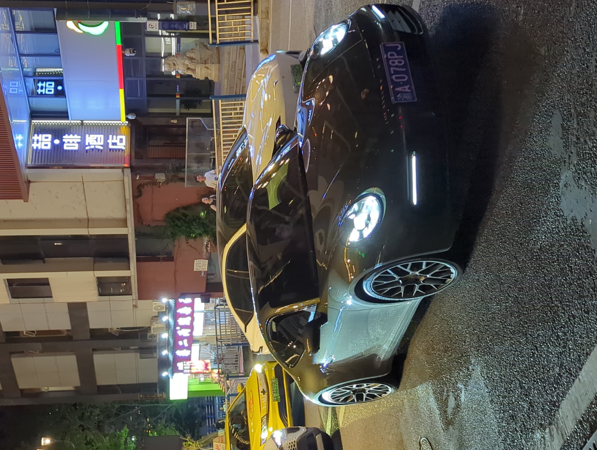 The image shows a nighttime street scene in Yuzhong District, China. In the foreground, a dark-colored Porsche sports car is parked or slowly moving on a wet asphalt road. The car's headlights and daytime running lights are illuminated. A license plate with the characters "A 078PJ" is visible on the front of the Porsche. Behind the Porsche, a white luxury sedan is also visible, with its headlights on. Further in the background, a yellow taxi with illuminated headlights and a license plate starting with "A A03563" is present. The scene is bordered by buildings, one of which has illuminated signage in Chinese characters. Two individuals, a woman and a man, are standing near the building in the background, looking in the same direction. The overall environment suggests a commercial or entertainment district at night.