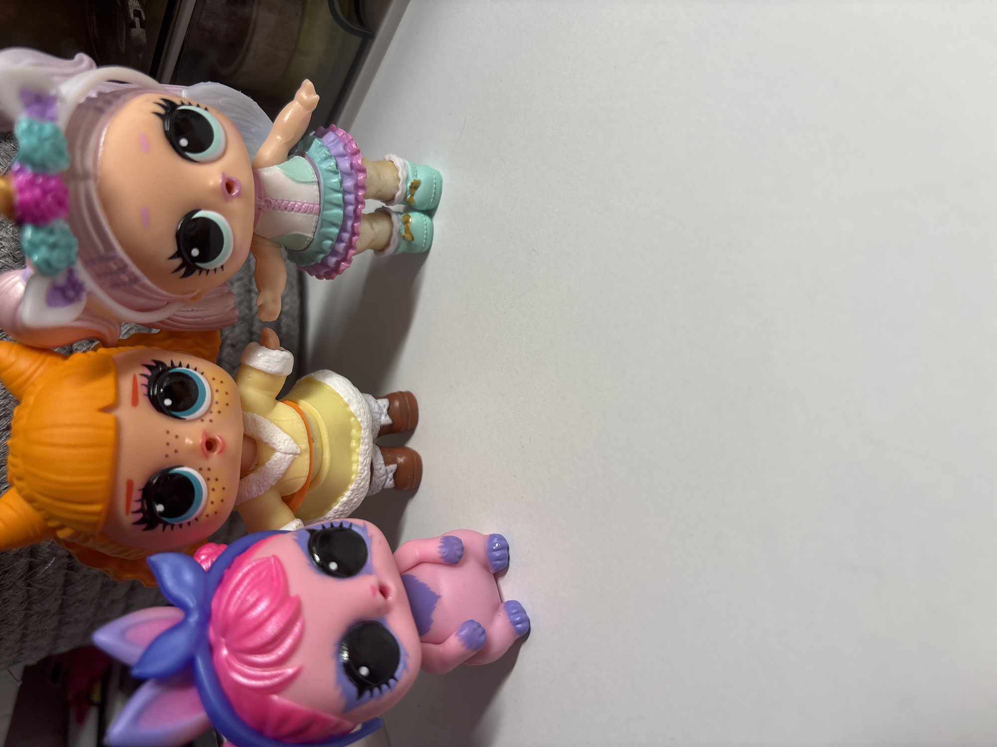 Three LOL Surprise dolls are posed on a white surface. From left to right, the first doll has pink hair styled with blue bunny ears and a purple bow. It has a pink body with purple markings on its face and chest, and black eyes. The second doll has orange hair and is wearing a yellow outfit with a white furry trim. It has large blue eyes and a light complexion with freckles. The third doll is wearing a unicorn-themed outfit with a white unicorn horn headband. It has light pink hair and a multi-colored skirt. The dolls are positioned in a way that suggests they are standing. The background is blurred, but appears to include a basket and some other objects. There is no indication of the specific location in the city of Velbert, Germany.