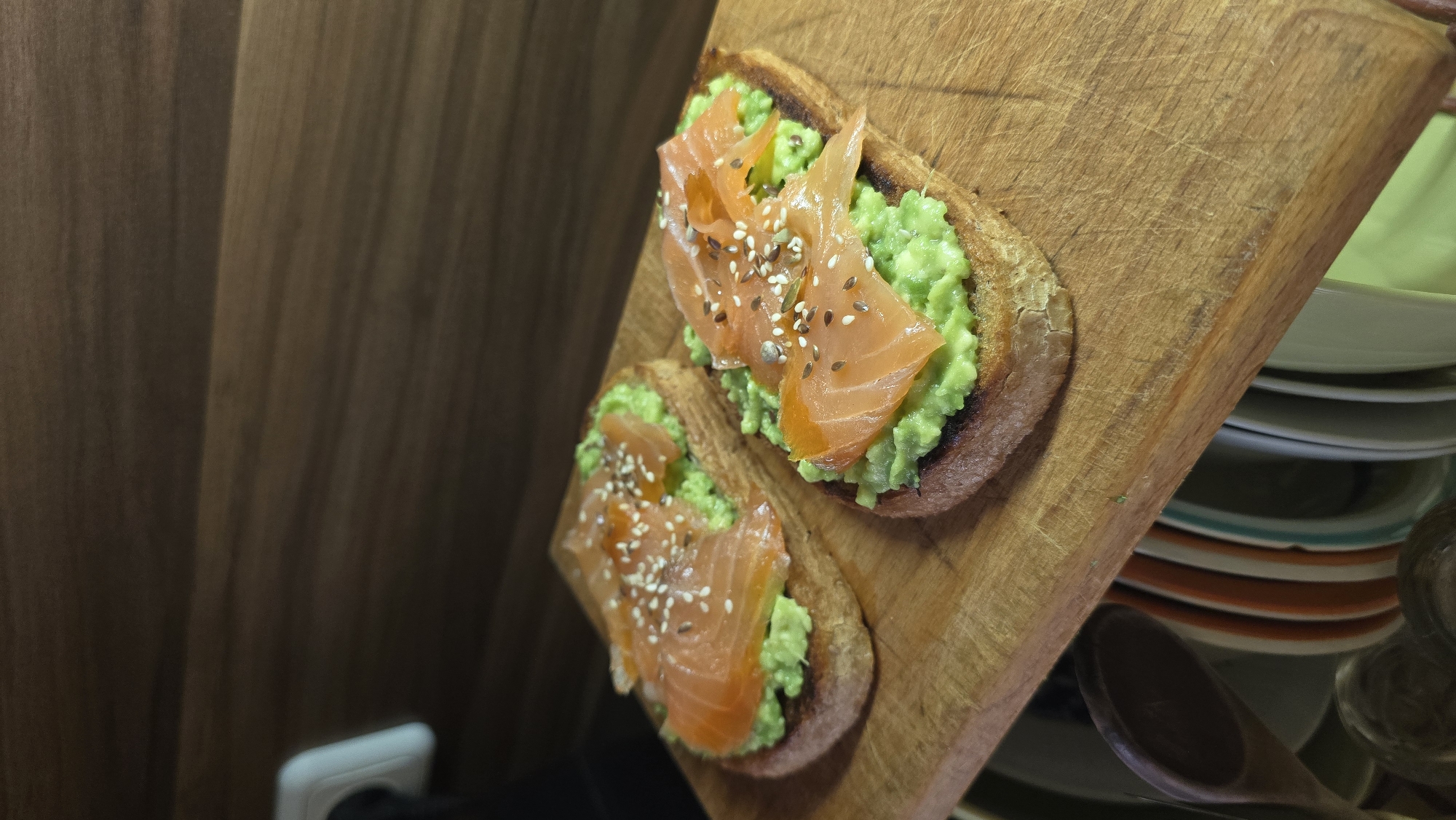 Two pieces of avocado toast topped with salmon and seeds are placed on a wooden cutting board. The wooden cutting board is positioned on a countertop, with a wooden wall behind it. To the right of the cutting board is a stack of plates and a wooden spoon. A white electrical outlet is visible in the bottom left corner of the image. The image does not contain any information regarding Stara Zagora, Bulgaria.