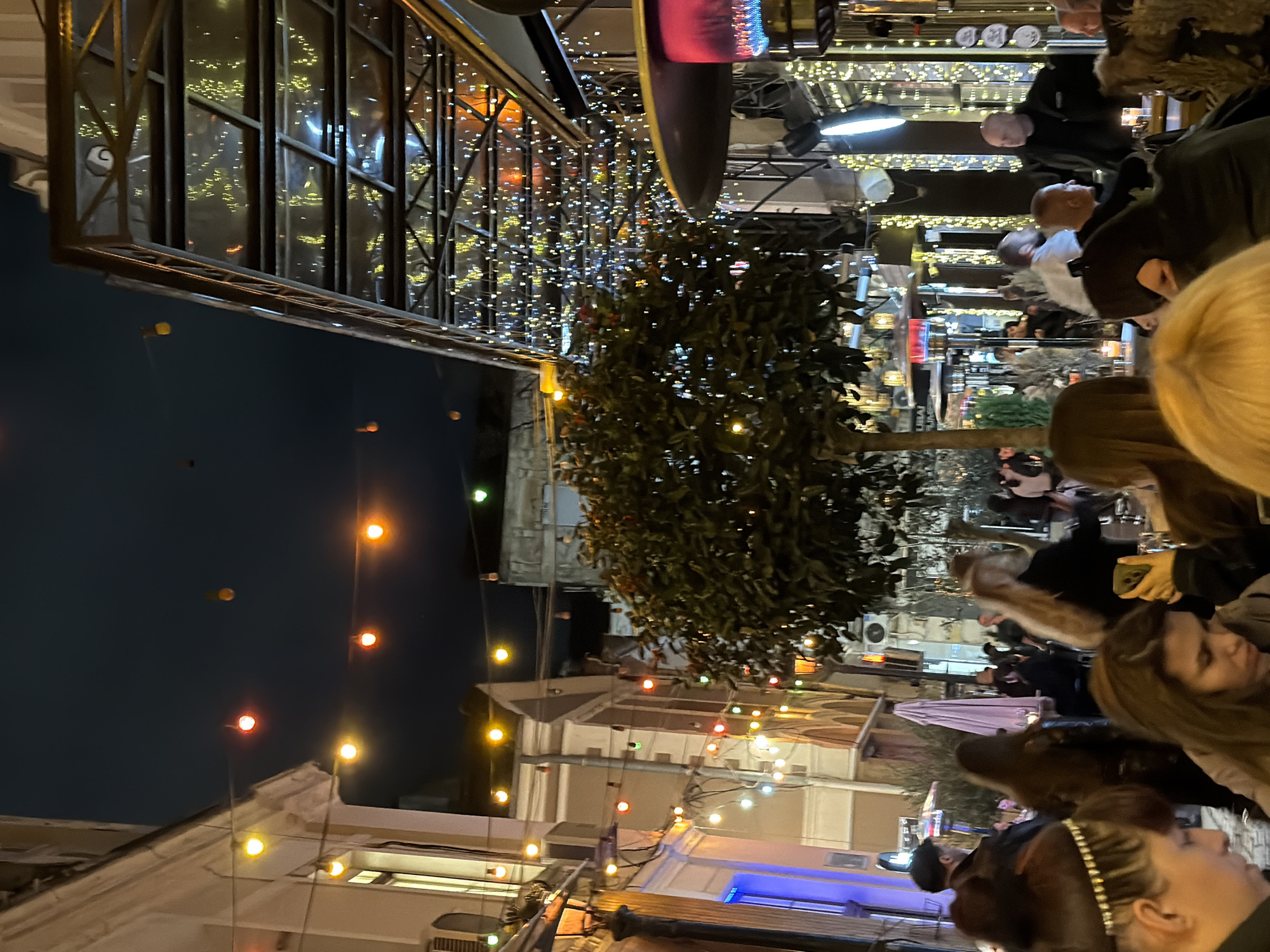 The image depicts an outdoor dining area in Plovdiv, Bulgaria, at night. The scene is brightly lit with various string lights and spotlights. There are several people seated at tables, appearing to be engaged in conversation and dining. The perspective suggests the image was taken from a relatively low angle, looking upwards towards the building and dining area. Buildings in the background are visible, adorned with string lights, adding to the ambiance. There are trees decorated with lights that give the foreground texture.
