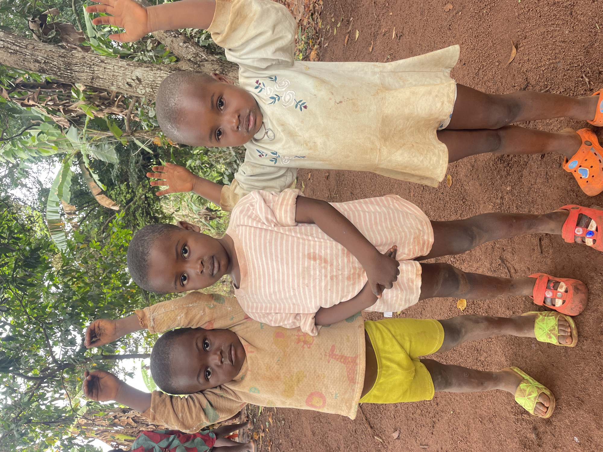 Three young children are lying in a line on a red dirt surface. The child on top wears a light yellow dress with floral embroidery and orange sandals. The middle child wears a pink and white striped shirt and red sandals. The bottom child wears a yellow shirt and yellow shorts and green sandals. All three children are looking at the camera. The background contains a tree and lush green vegetation. This image was taken in Ogbe, Nigeria.