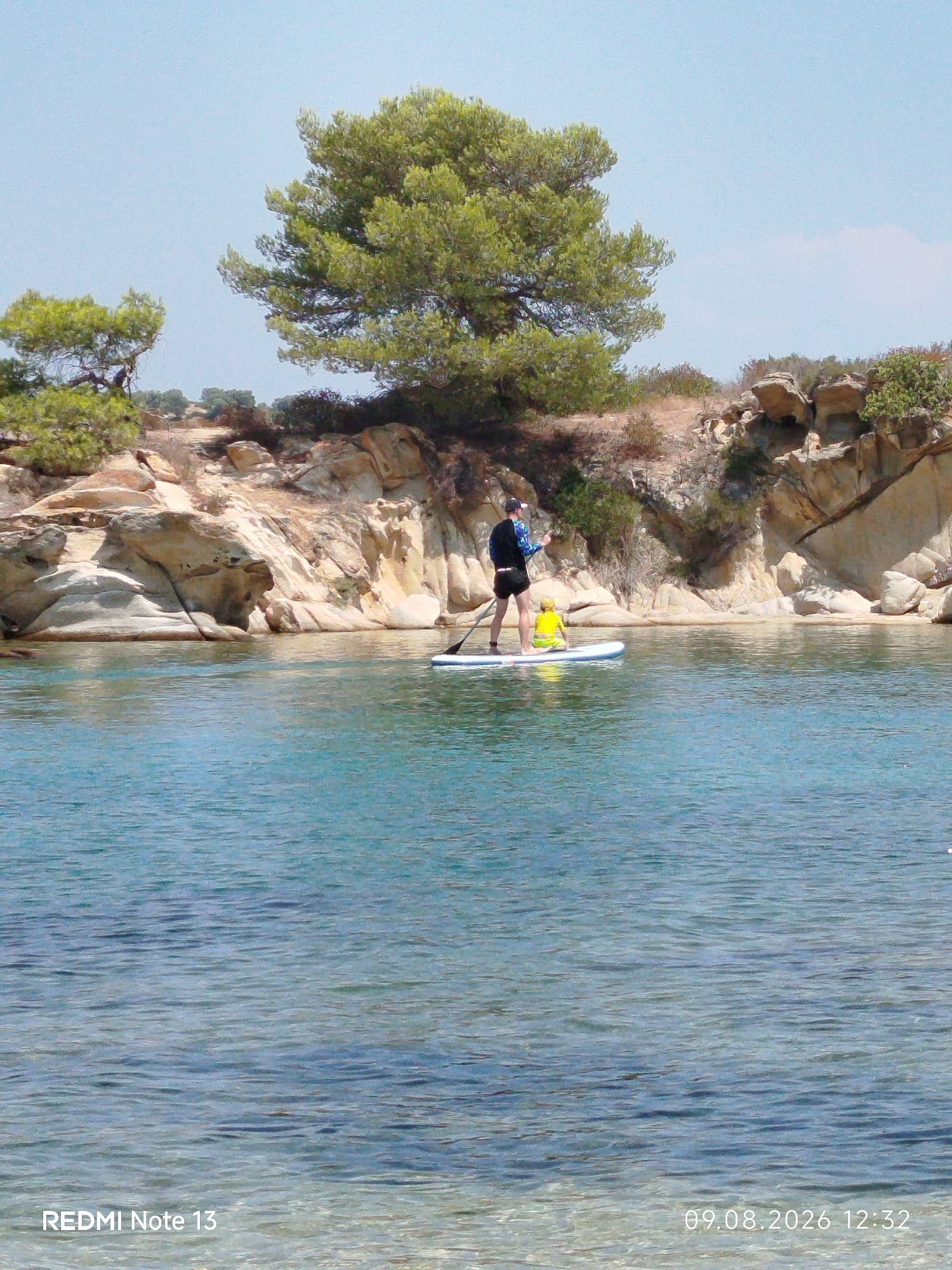 Two people are on a paddleboard in the clear, blue water. One person, standing and holding a paddle, wears a black top and black shorts. The other person, a child, is sitting on the paddleboard wearing a yellow shirt. They are near a rocky coastline with sparse vegetation. The water is calm, reflecting the sky and the coastline. The image appears to be taken during the day under a clear sky. The timestamp in the bottom right corner indicates the date and time are 09.08.2026 12:32. The text "REDMI Note 13" is visible in the bottom left corner. This scene is located in Sithonia, a municipal unit in Greece, known for its picturesque beaches and clear waters.