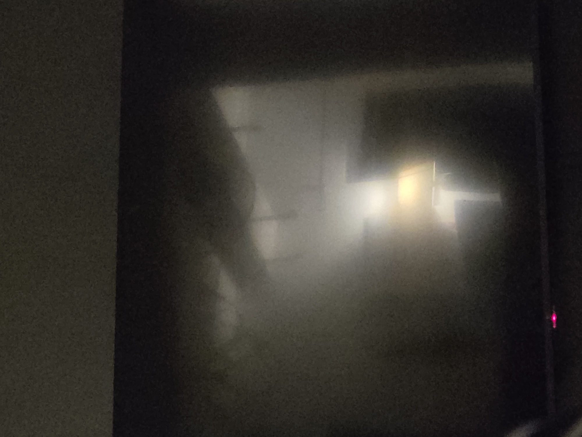 The image shows a reflection on a dark surface, likely a screen or a window. In the reflection, a lamp with a beige shade is visible, emitting a warm light. Behind the lamp, there is a blurred background that suggests an indoor environment with some furniture. In the lower center of the image, a small red light is visible on the dark surface itself. The scene is dimly lit and appears to be taken at night. There is no indication of the city of Voluntari, Romania, or any specific location.