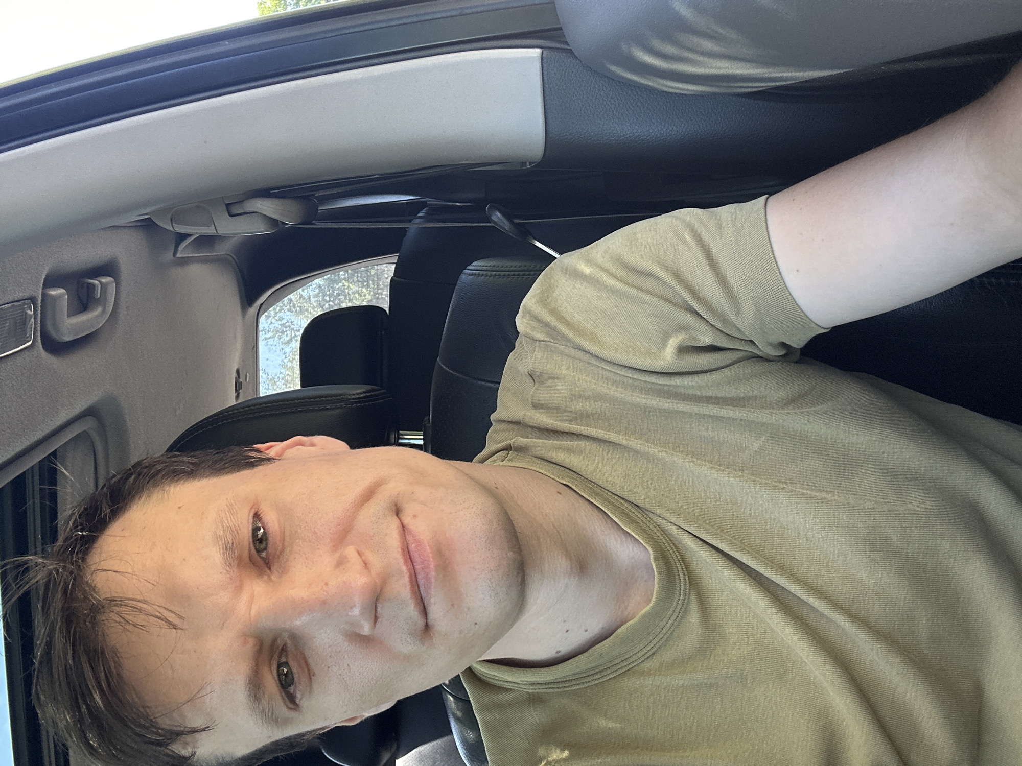 A man is taking a selfie inside a car. He is wearing a tan t-shirt and has short brown hair. The interior of the car is visible, including black leather seats, the ceiling, and a sunroof. The man appears to be in the driver's seat, and the photo is taken from a slightly high angle, looking down at him. The scene is well-lit, suggesting it is daytime. There is no specific indication of the location within the city of Makiv, Ukraine, beyond the fact that the man is inside a vehicle.