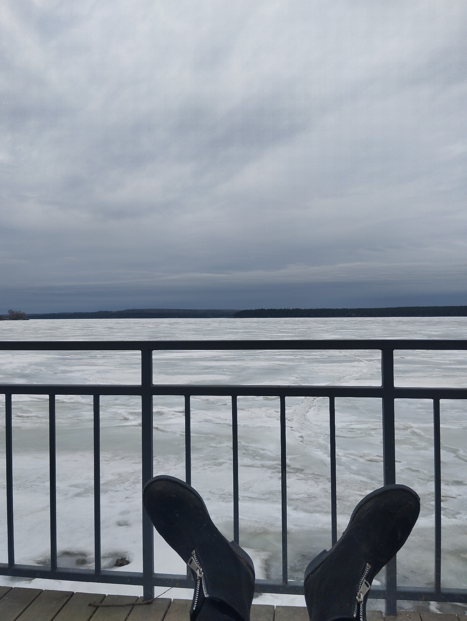 The image shows a first-person view of someone sitting on a wooden deck or pier, with their feet stretched out towards a metal railing. Beyond the railing is a partially frozen body of water, likely a lake or wide river, covered with ice floes and patches of open water. In the distance is a treeline. The sky is overcast with gray clouds. The person is wearing black boots with visible silver zippers on the sides. Given the location in Votkinsk, Russia, it is likely that the body of water is the Votkinsk Reservoir.