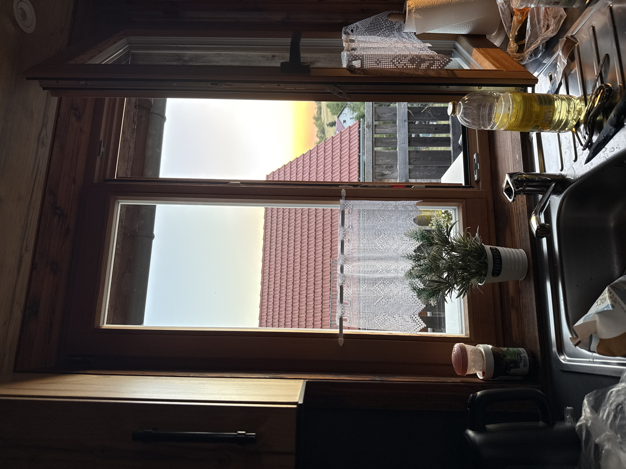The image depicts a kitchen window with a view of a rural landscape, likely in Breungeshain, Germany, given the architectural style of the house roofs. The window is open, revealing a warm sunset with a clear sky. Inside the kitchen, a sink is visible in the foreground, with a faucet, a bottle of cooking oil, a knife, and other kitchen items. A potted plant sits on the windowsill, and a white lace curtain hangs across the lower part of the window. The interior of the kitchen appears rustic, with wooden elements and warm lighting. The overall scene suggests a peaceful evening in a rural setting.