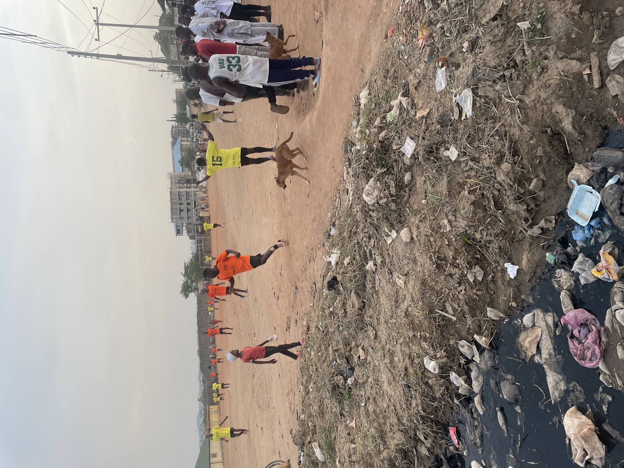 A dusty, brown, open field in Abuja, Nigeria, is the setting. Dozens of soccer players in orange and yellow training uniforms are lined up on the left side of the field, some jogging and stretching. Several spectators watch along the upper edge of the field. Two dogs trot in the middle ground. To the right, a large, trash-filled drainage ditch runs along the edge of the field, overflowing with plastic waste, stagnant water, and general debris. Power lines are visible in the upper left of the frame. A partially constructed building is behind the players.