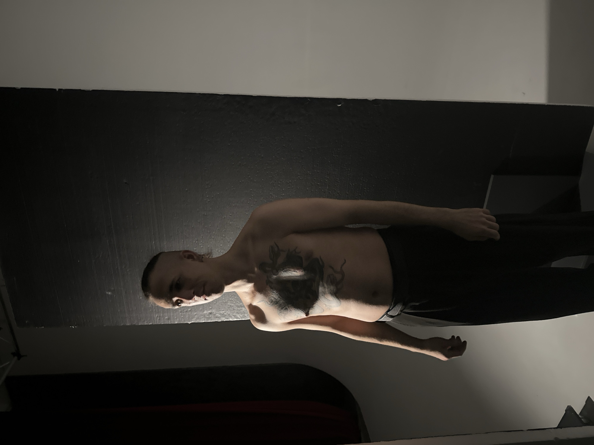 The image displays a male individual standing indoors, reportedly in Saratov, Russia. The man, appearing to be in his late teens or twenties, is bare-chested, revealing a prominent dark tattoo covering his central chest, possibly depicting a figure, and a smaller tattoo below his left pectoral muscle. He has short hair on the sides with a longer top, styled back. He wears dark-colored trousers with a dark belt. He stands upright, facing generally forward but with his head turned slightly to his left (viewer's right), his gaze directed off-camera. His arms are relaxed at his sides. The environment consists of a stark background: a light gray wall on the right side and a dark, textured, possibly black wall or partition on the left. A dark, cylindrical or bag-like object is partially visible on the far left of the frame, against the light gray wall. The scene is lit by a strong, directional light source from the viewer's left, highlighting the subject's front and right side and casting shadows to his left. No other individuals or direct interactions are depicted.