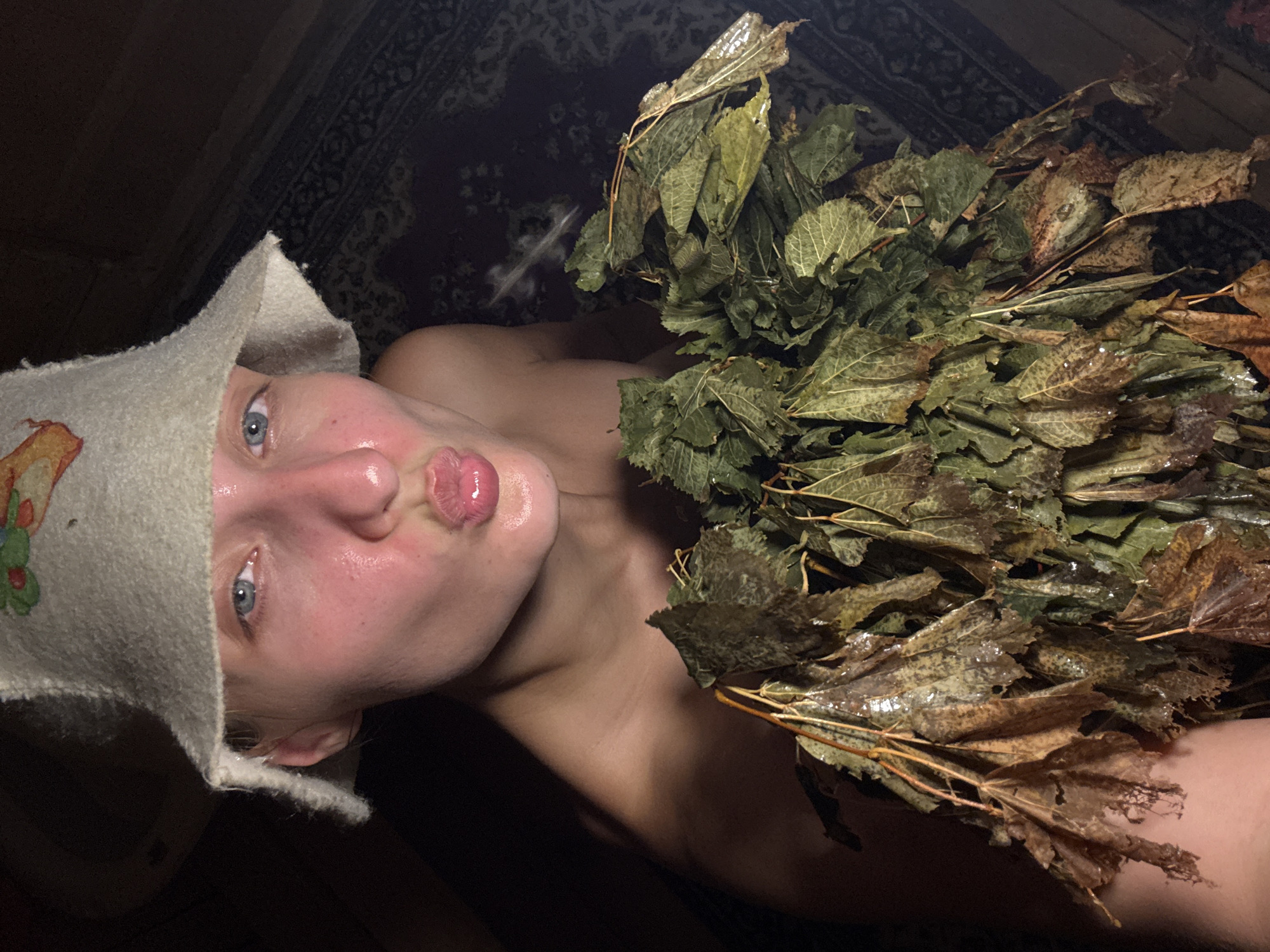 In the image, a young person is shown wearing a white felt hat with a colorful embroidered image of an animal. They have blue eyes and pink, sweaty skin, with their lips pursed as if blowing a kiss. They are holding a bunch of wet, dried leaves and branches in front of their chest. The background appears to be a dimly lit room with dark wooden walls and a patterned rug on the floor. The scene suggests a sauna or a similar steamy environment.