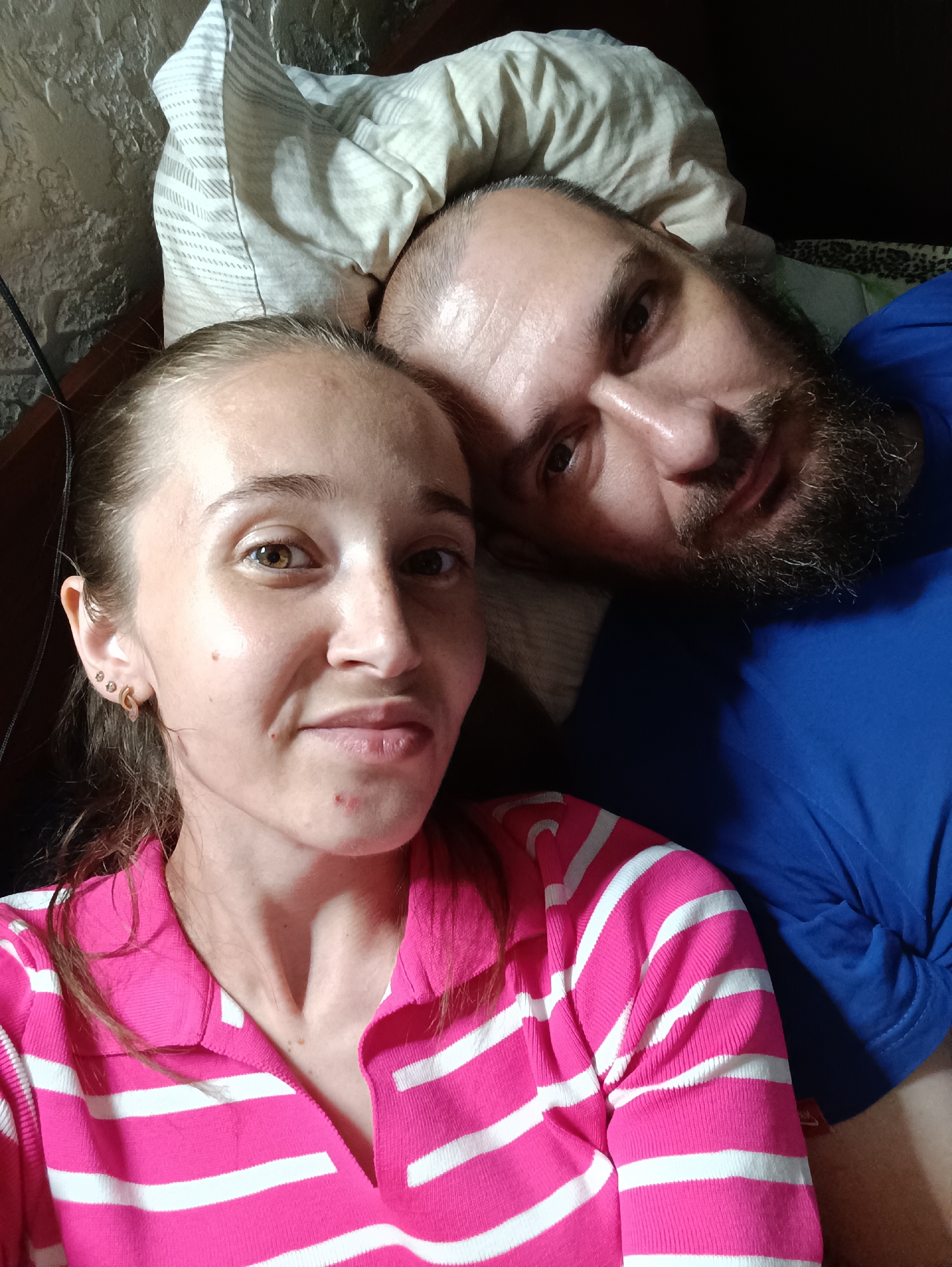 In Vypasne, Ukraine, a close-up, over-the-shoulder selfie is taken from a low angle, looking up at two individuals lying down, possibly in a bed. The person in the foreground, on the left, is a woman with light brown hair pulled back. She has fair skin, brown eyes, and is wearing a pink and white striped polo shirt. She has three small earrings in her left ear. To the right and slightly behind her, a man with dark hair and a beard is visible. He is wearing a blue t-shirt and his head is resting near the woman's. The background shows a pillow with a striped pattern and a dark, textured surface that may be a wall.
