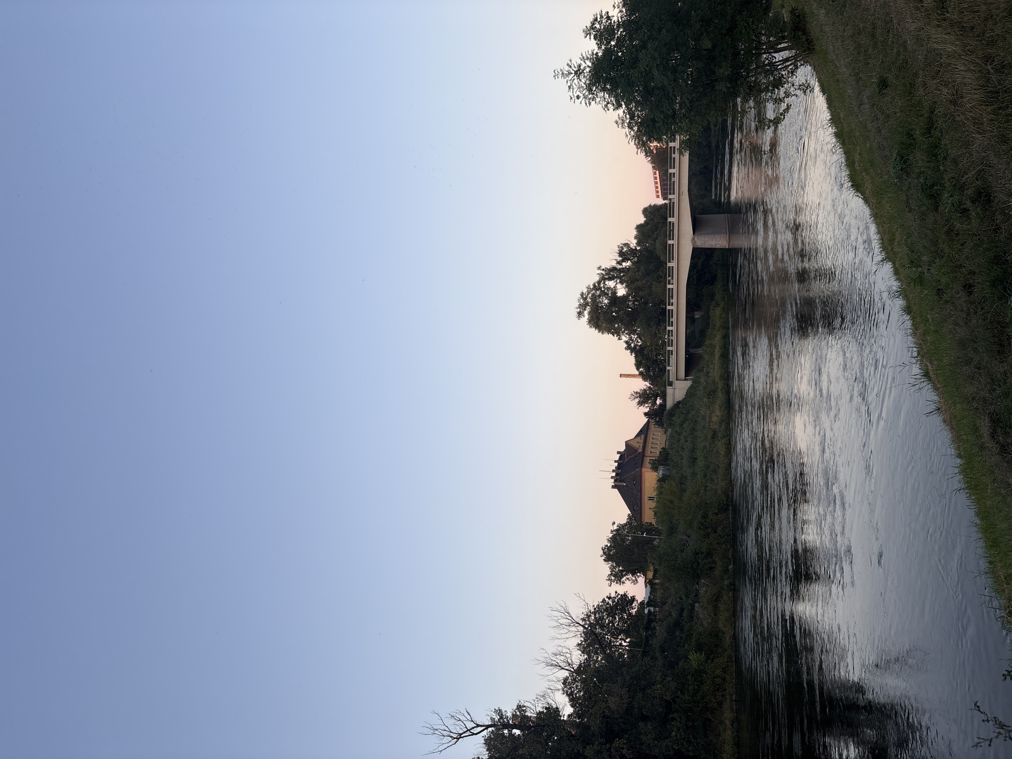 A wide, dark river flows from left to right across the foreground, its surface rippling. The bank on the right is covered in grass and a small tree. On the left bank, dense green trees and some bare branches create a dark silhouette against a pale, rosy sky. In the middle distance, a light-colored bridge spans the river, supported by concrete pillars. Beyond the bridge, a yellow building with a dark roof is visible amongst more trees. The sky above is a gradient of pale blue, transitioning to a soft pink near the horizon.