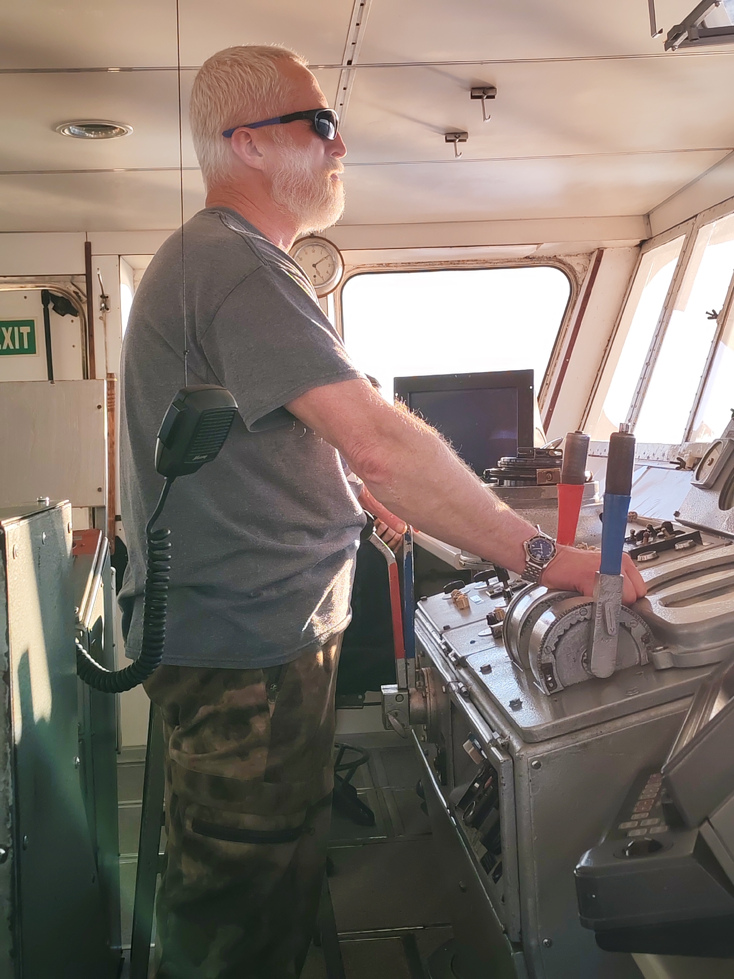 A middle-aged man with gray hair and a beard stands at the helm of a boat, likely a ferry or small cargo ship, in Ozernoe Ust'e, Russia. He is wearing sunglasses, a gray t-shirt, and camouflage pants. A coiled microphone is attached to his shirt. He has his hands on the controls of the boat. A monitor, clock and exit sign are also visible. The environment looks to be a bright, sunny day.