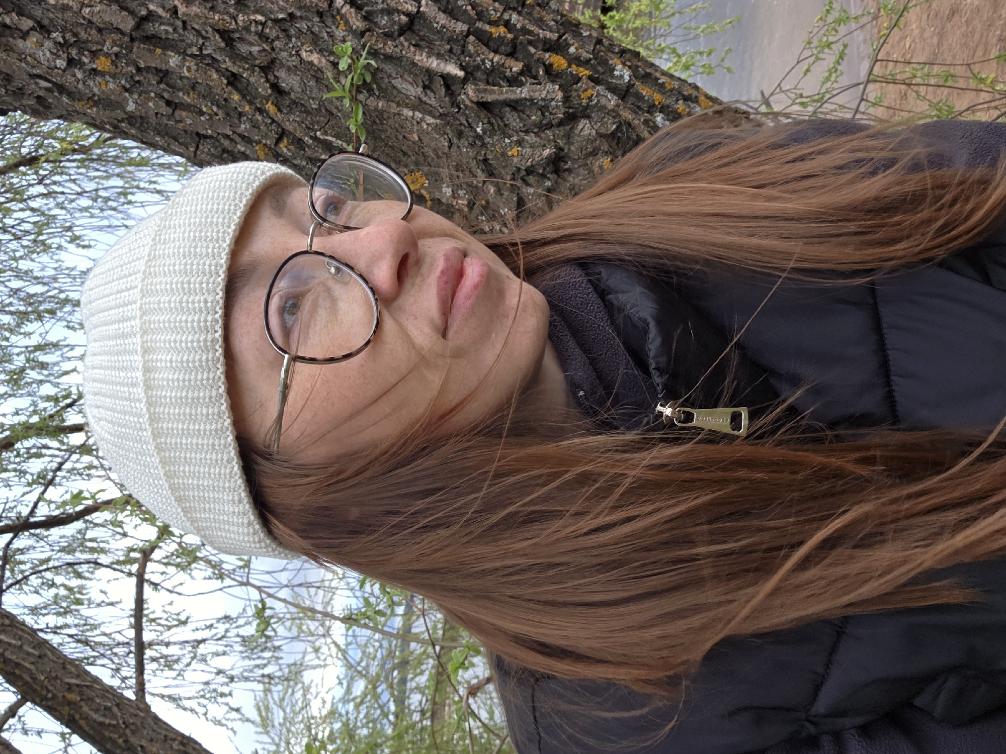 A woman wearing a white beanie, round glasses, and a black puffer jacket is positioned against a tree. Her long, brown hair is loose. She is looking upwards. In the background, branches and green leaves are visible, suggesting it is outdoors, possibly in a park. The image was taken in the city of Chaikovsky, Russia.