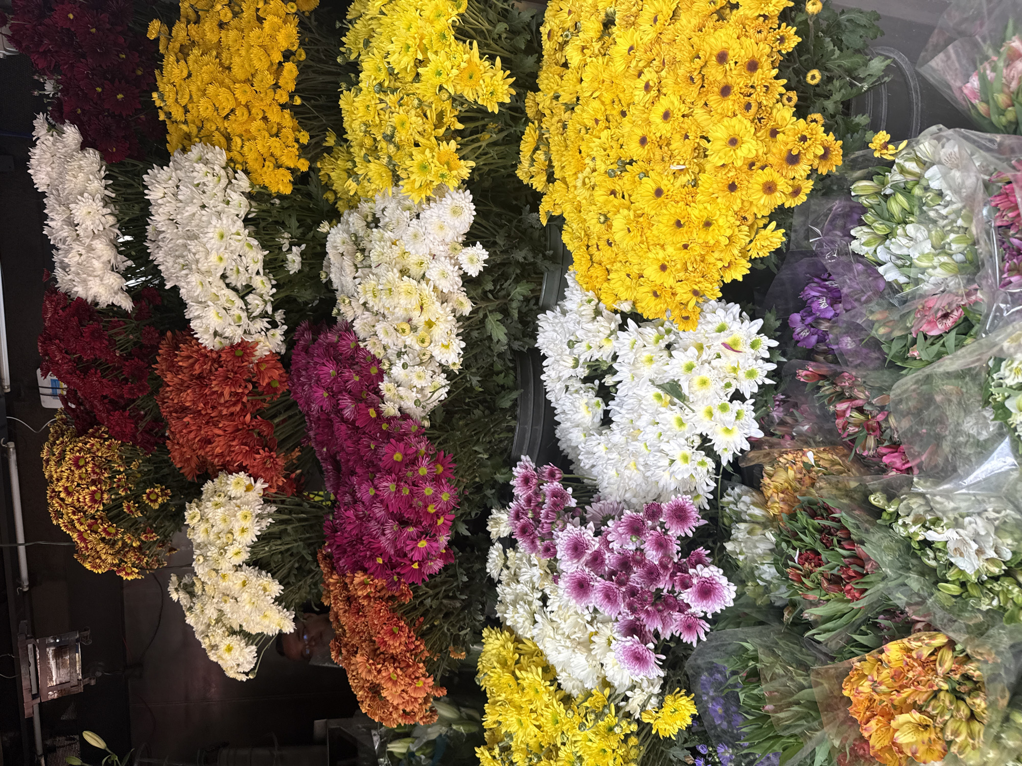 The image shows a high-angle view of various flower arrangements, likely taken in Mexico City, Mexico. The flowers are primarily chrysanthemums and alstroemerias in a wide range of colors, including yellow, white, red, orange, and purple. The flowers are arranged in bunches, with some wrapped in plastic. The scene likely takes place in a flower market or a flower shop.