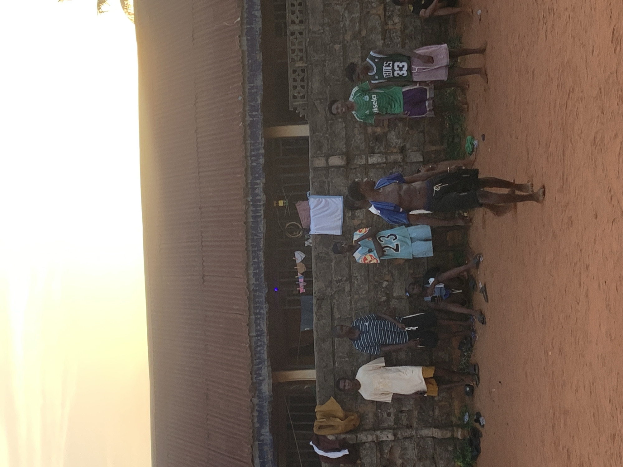 In Ukwu-Abwa, Nigeria, a group of people are gathered outdoors, likely near a building with a corrugated metal roof and a brick or stone wall. Some of the individuals are wearing jerseys, while others are in casual attire. Laundry is hanging outside a window. The ground appears to be dirt or sand. The light suggests it may be early morning or late afternoon.