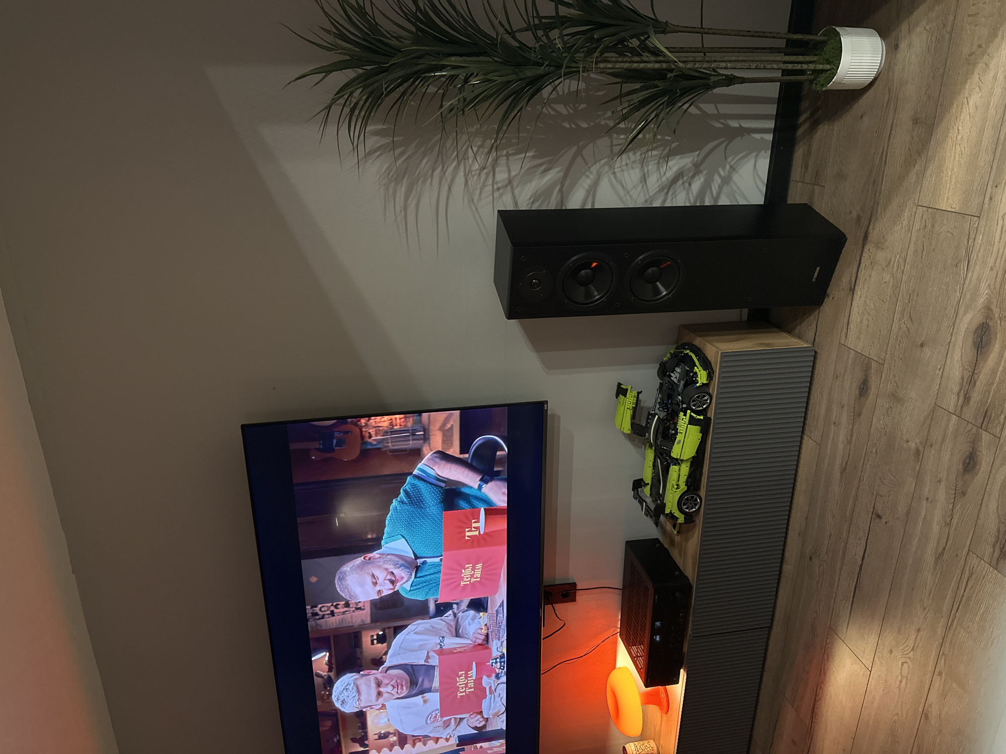 The image shows a living room interior. On the wall is a flat-screen television displaying a scene of two people reading boxes labeled in Cyrillic, possibly in a televised production. Below the TV is a console, with audio equipment, and a desk lamp on the console. The wall also features a central speaker. A toy car sits atop the console and there is a potted plant in the corner of the wall. The general interior design suggests a contemporary style. The image may have been captured in a location within the city of Perm, Russia, suggested by the Cyrillic text on the television.