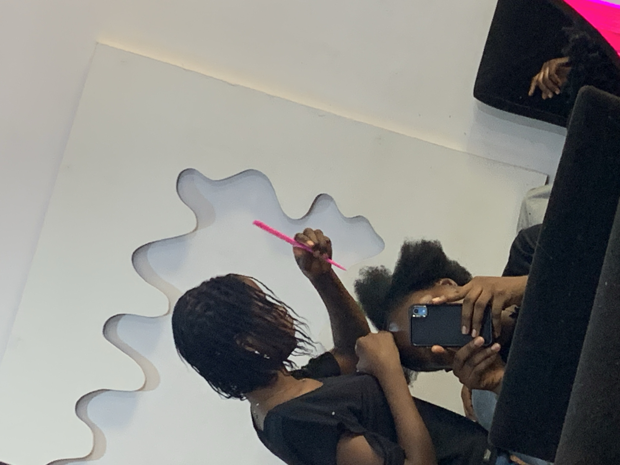 The image depicts two individuals engaged in hairstyling. One person is holding a pink comb near another person's head, likely styling their hair. The second person is holding a smartphone, presumably taking a picture or recording the process. The scene appears to be indoors, with a white wall and a decorative cutout behind them. The setting of Jos, Nigeria cannot be confirmed from this image.